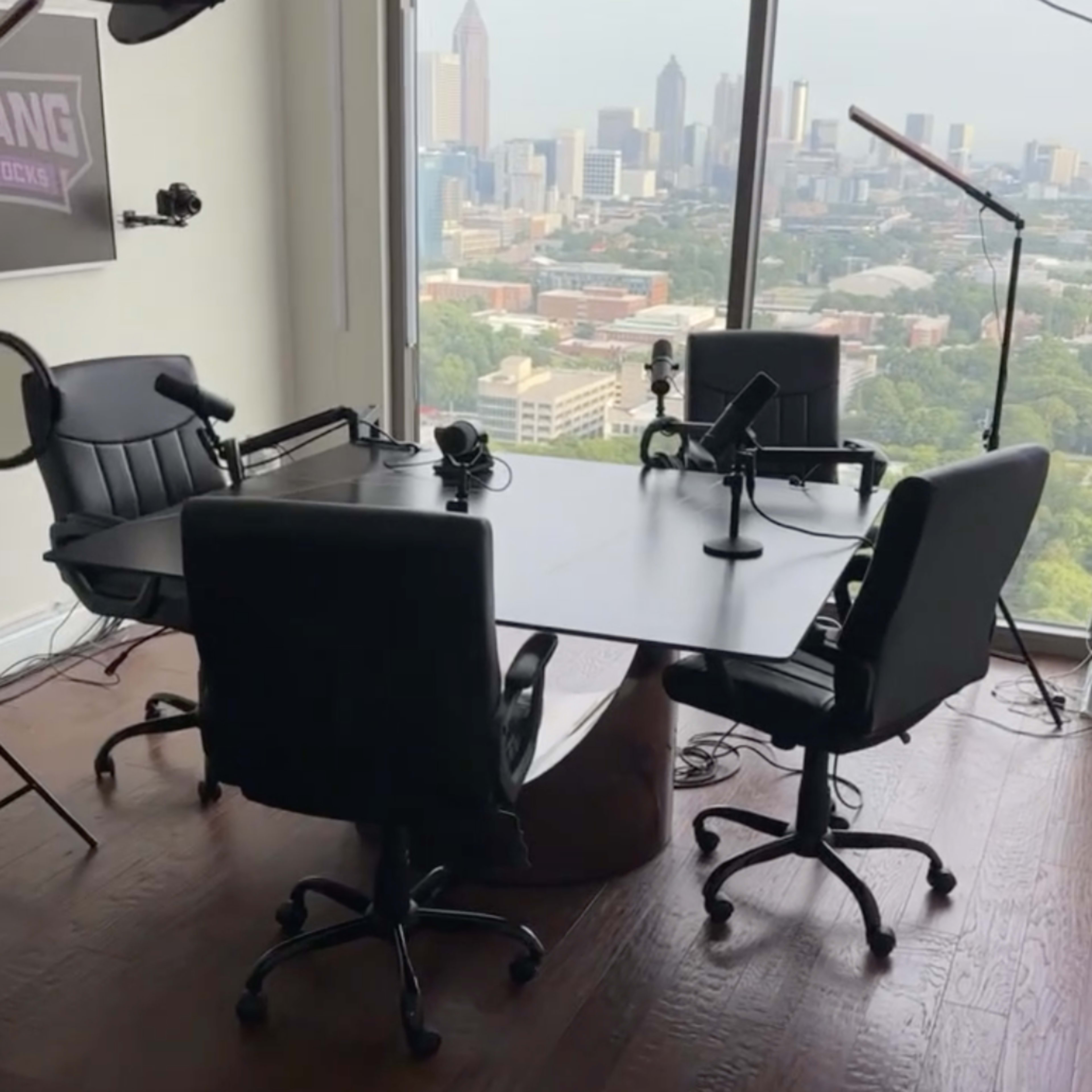 A modern podcast studio with a round table, four black chairs, microphones, and large windows offering a view of a city skyline.