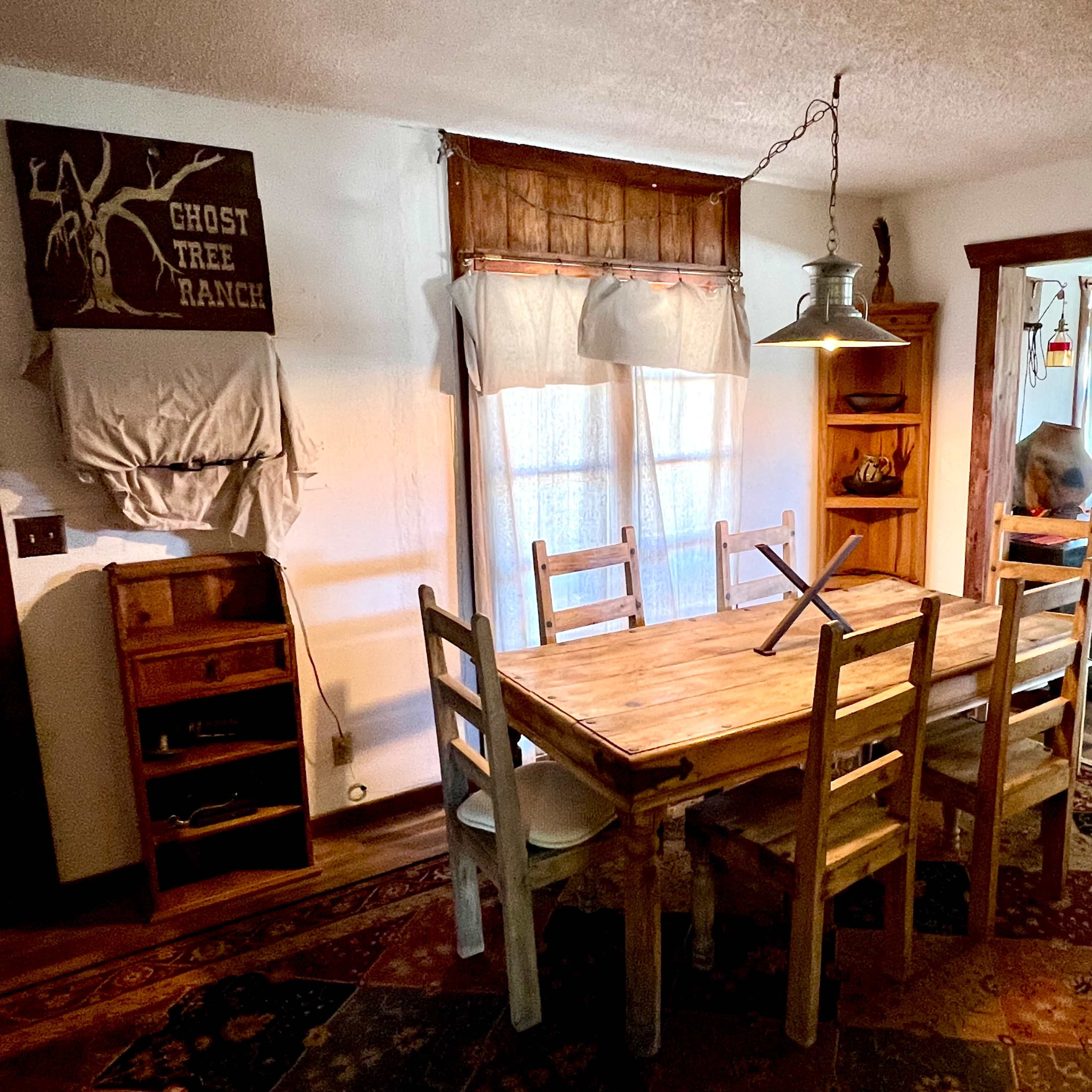 A rustic dining area features a wooden table surrounded by chairs, with a hanging light overhead and a view into a brightly lit adjacent room.