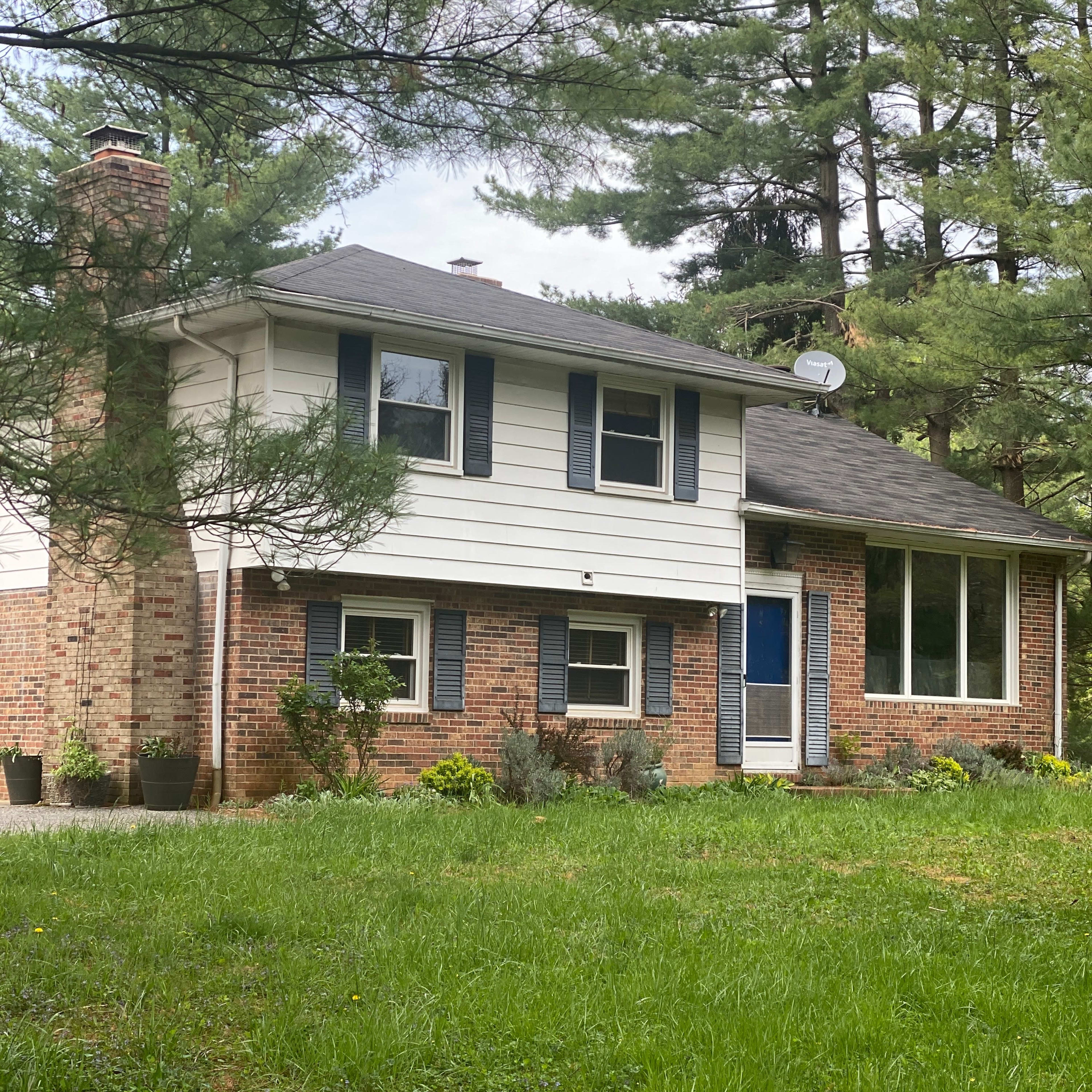 A two-story house with a brick and siding exterior is surrounded by tall pine trees and a grassy lawn.