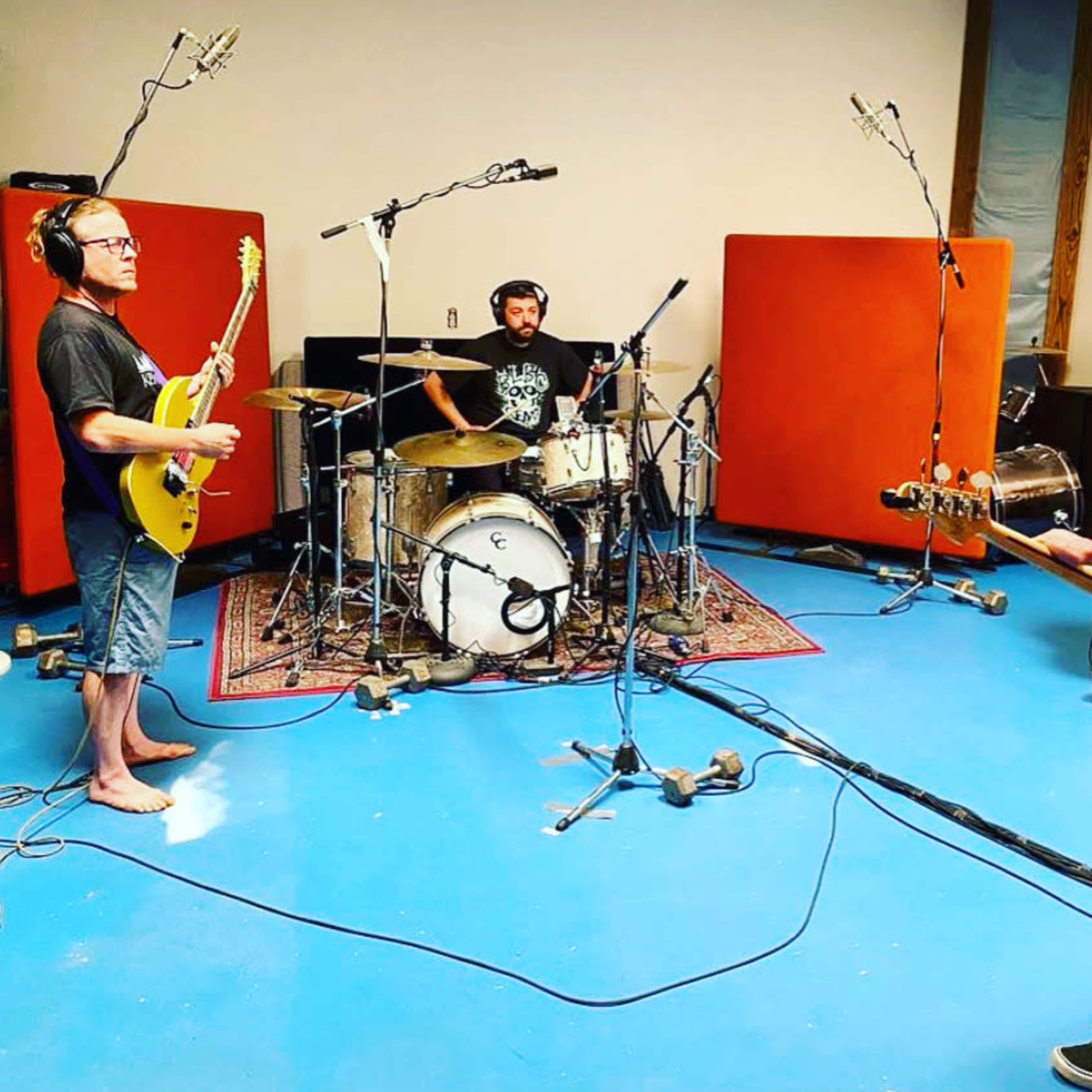 A group of four musicians is playing instruments in a sound studio with orange soundproofing panels and microphones set up around them.
