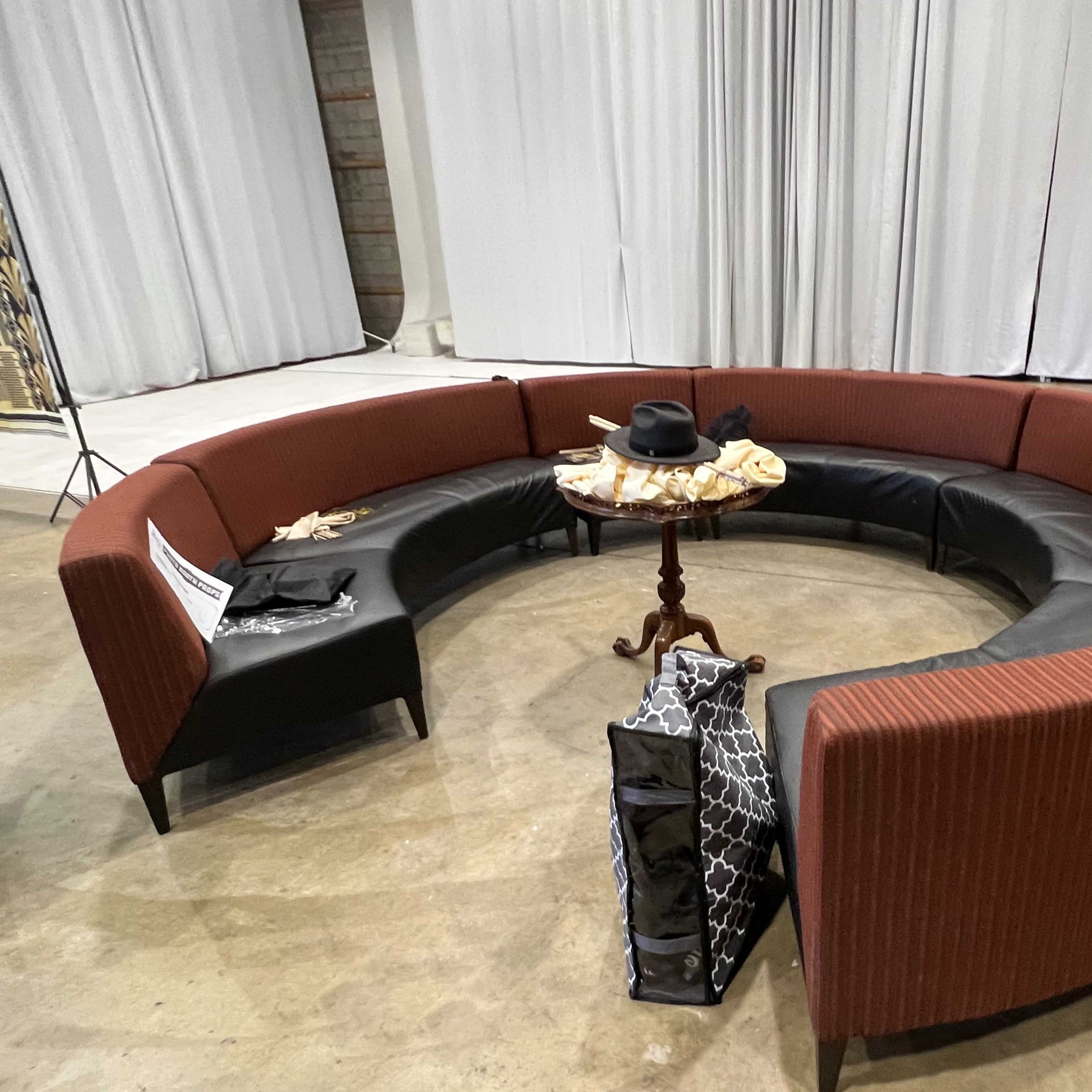 A circular seating arrangement with a dark red and black upholstered couch surrounding a small wooden table, placed on a concrete floor.