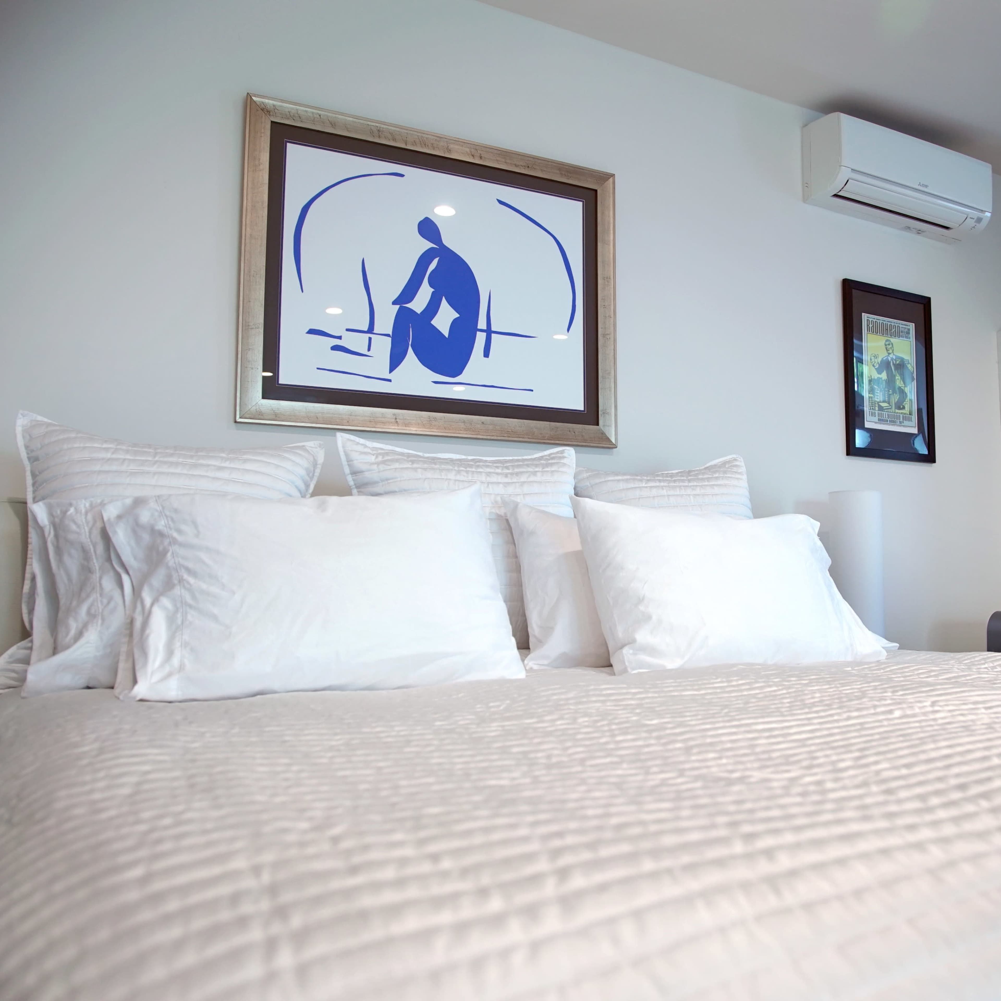 A neatly made bed with multiple white pillows is displayed in a modern bedroom featuring framed artwork and a compact kitchenette in the background.