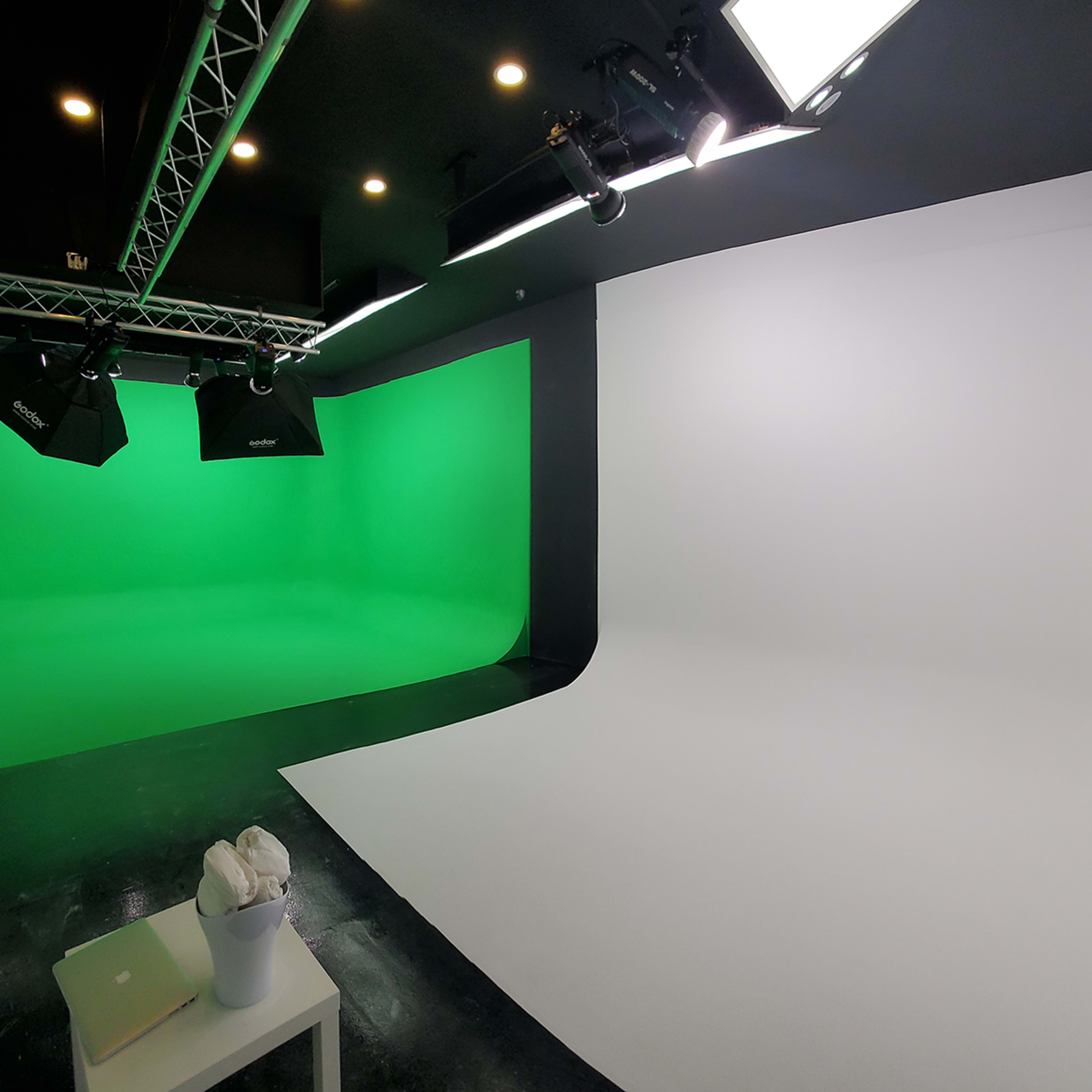 A studio space divided into two sections with a green backdrop on one side and a white backdrop on the other, along with lighting equipment and a white seating area.