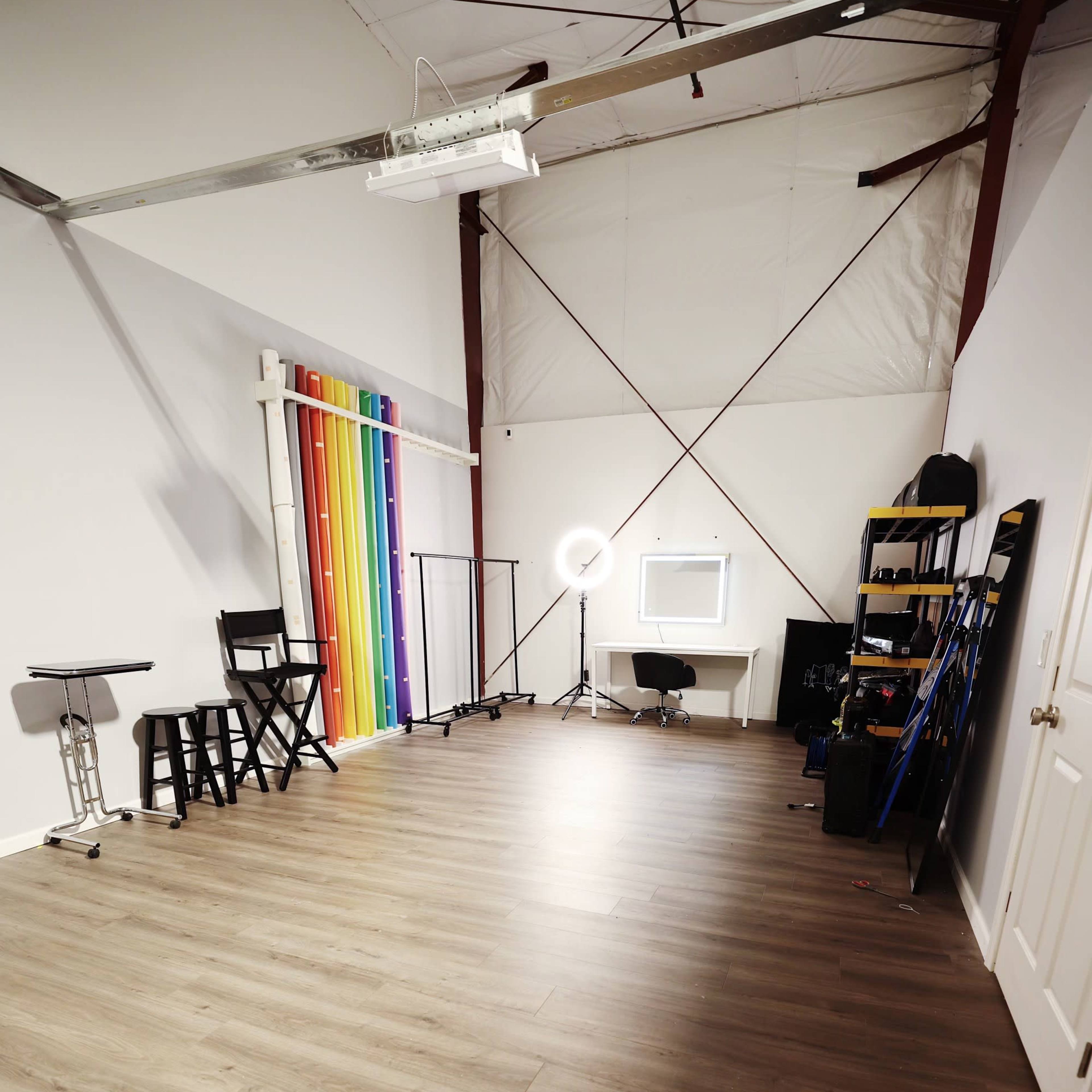 A spacious studio space with light wood flooring, a rainbow backdrop, a ring light, and various furniture items arranged along the walls.