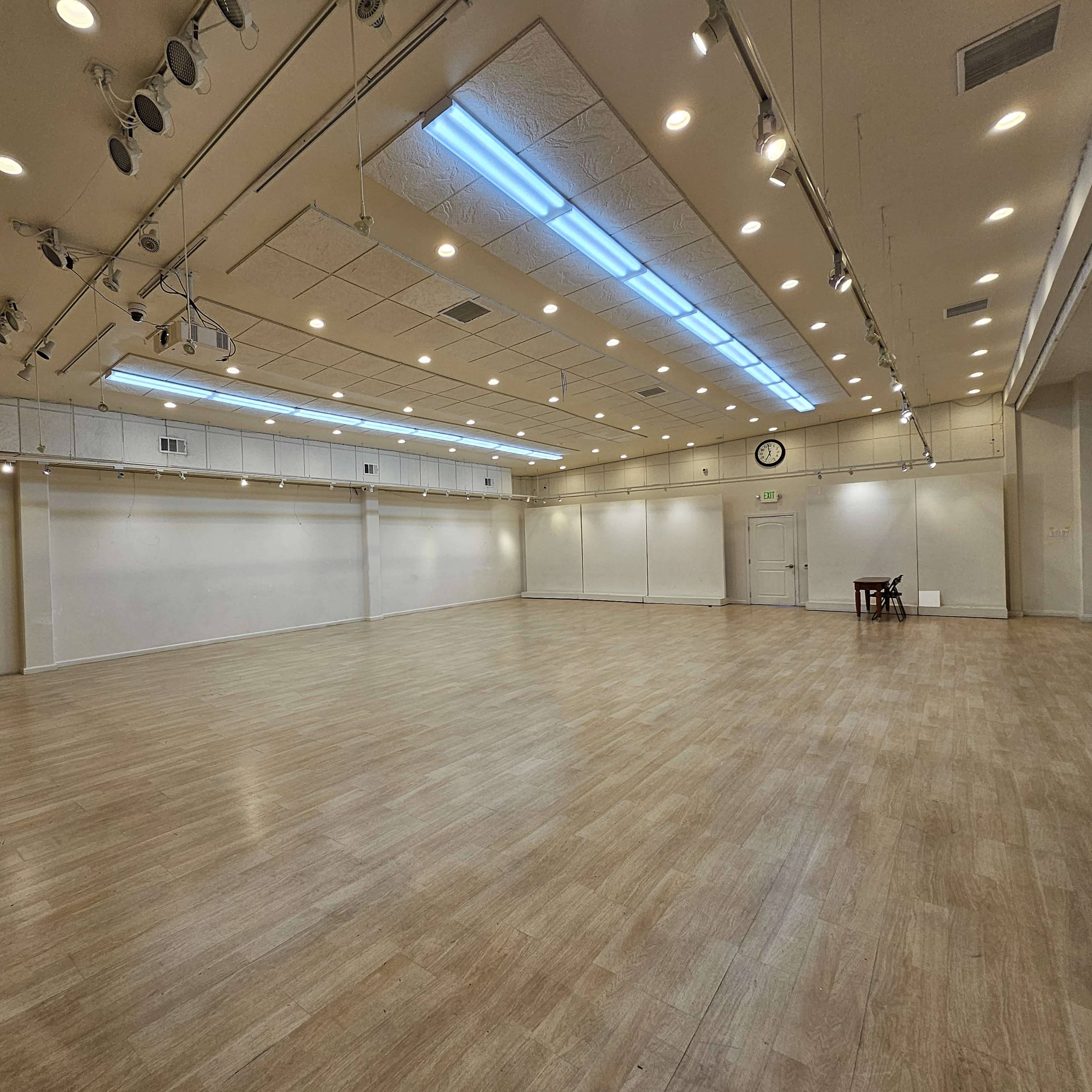 A spacious, well-lit room with wooden flooring and simple white walls, featuring a small table at one end.