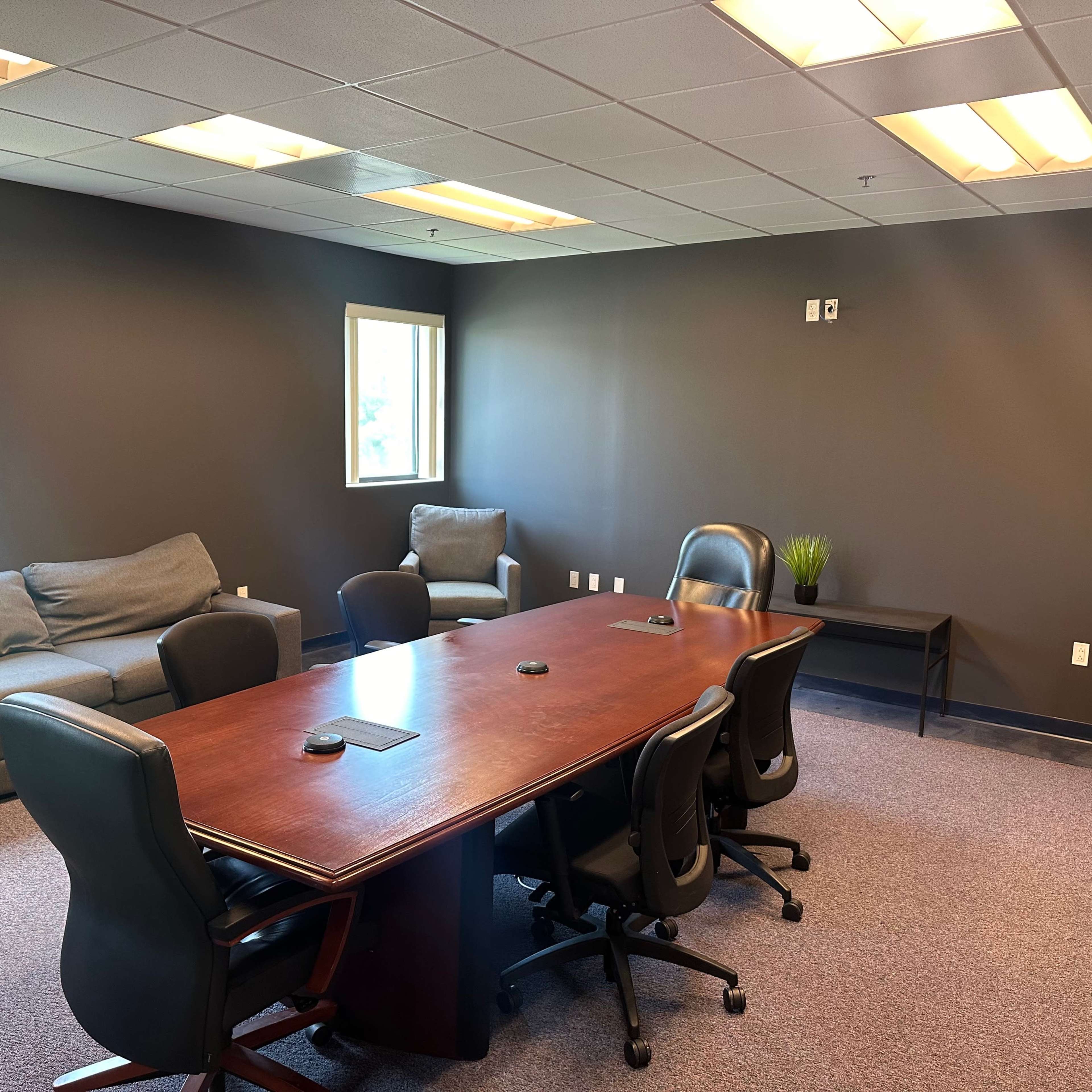 A conference room with a large wooden table surrounded by chairs, flanked by couches and a small side table against a dark wall.