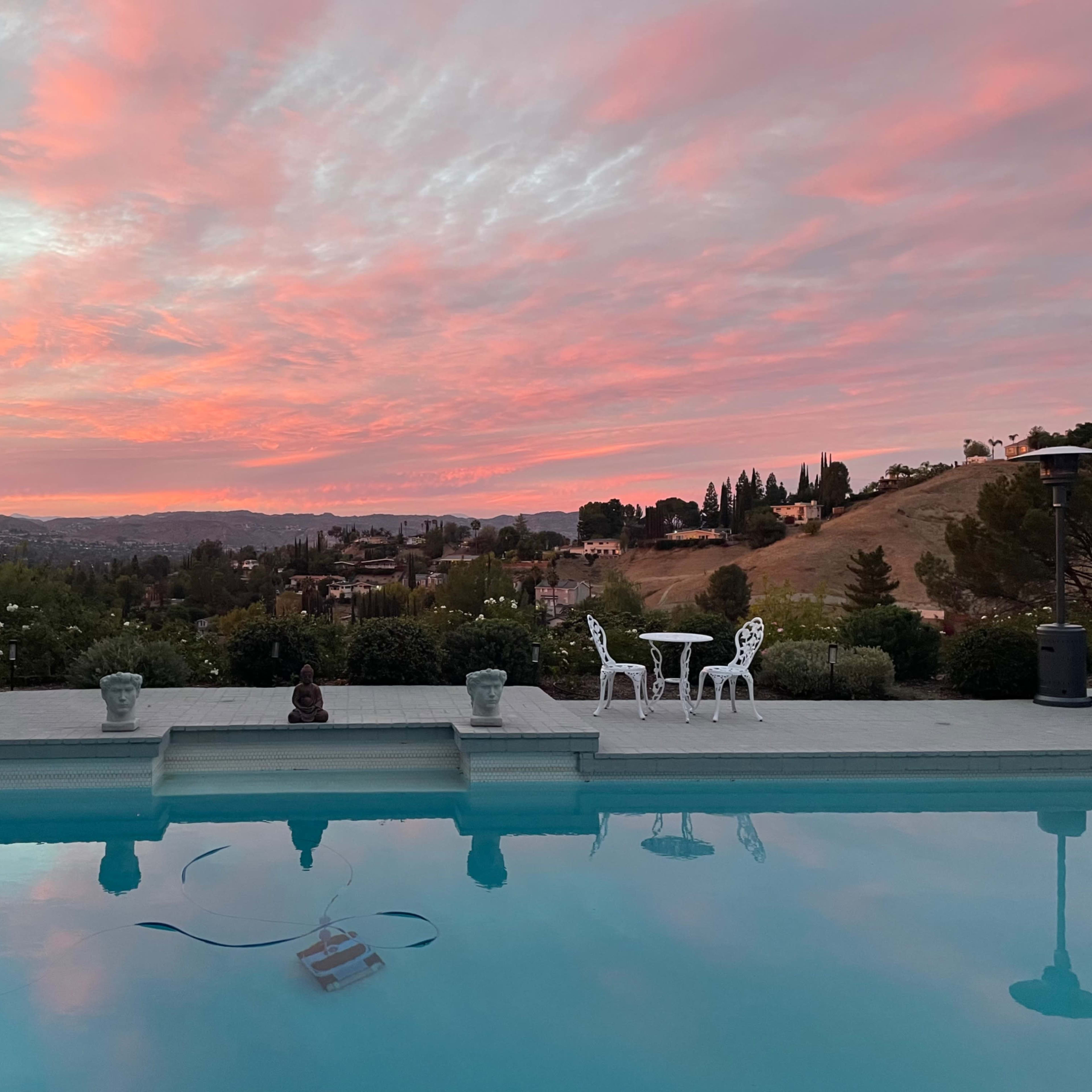 The scene features a swimming pool reflecting a colorful sunset sky, with a hillside view and two white chairs set near the water's edge.