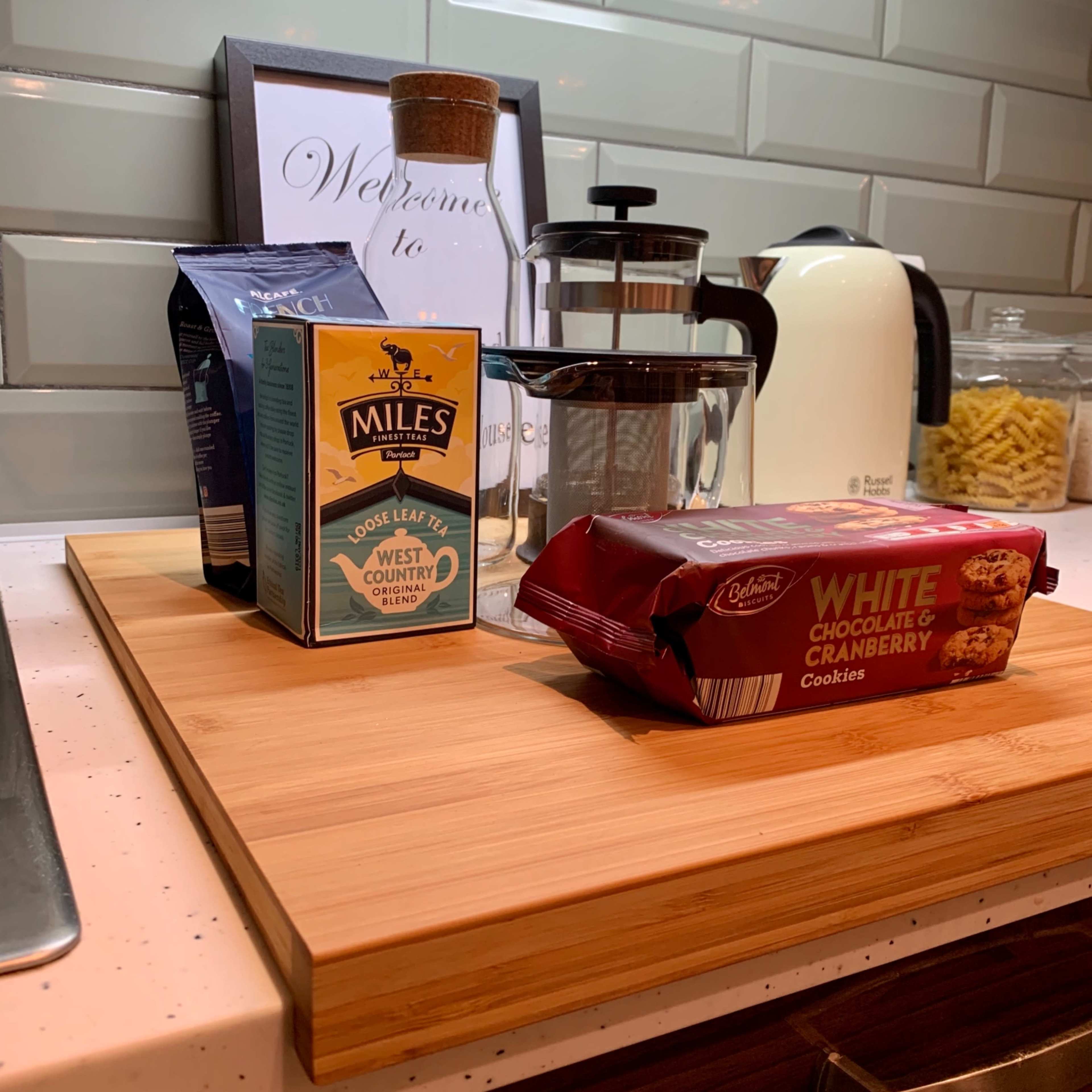 A bamboo cutting board with a box of tea, a French press, and a pack of white chocolate and cranberry cookies, set against a kitchen backdrop featuring a kettle and a jar of pasta.