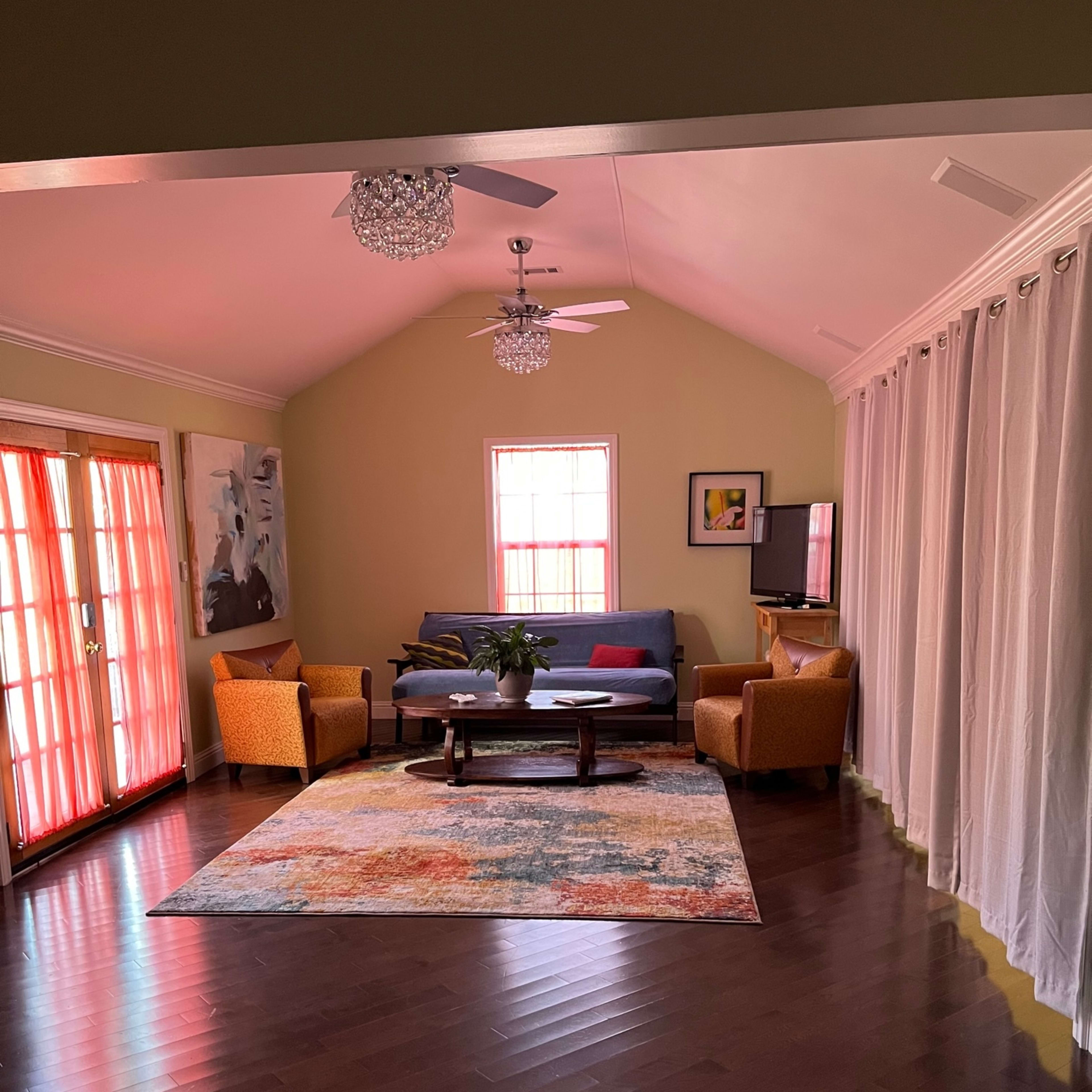 A bright living room with a blue sofa, orange armchairs, and a colorful area rug, illuminated by natural light filtering through sheer curtains and a glass door.