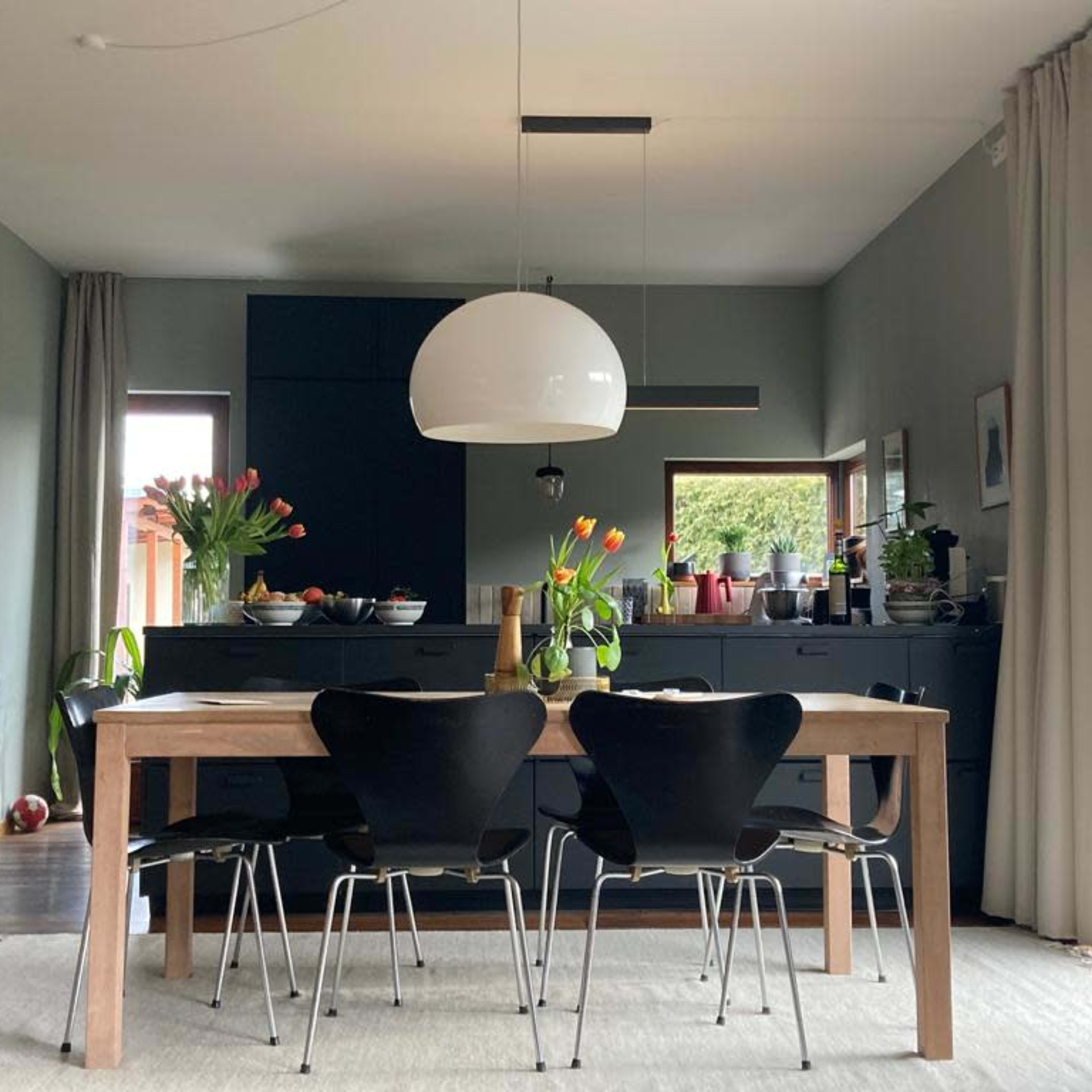 A dining room features a wooden table surrounded by black chairs, with a large pendant light above and a dark cabinetry wall in the background.