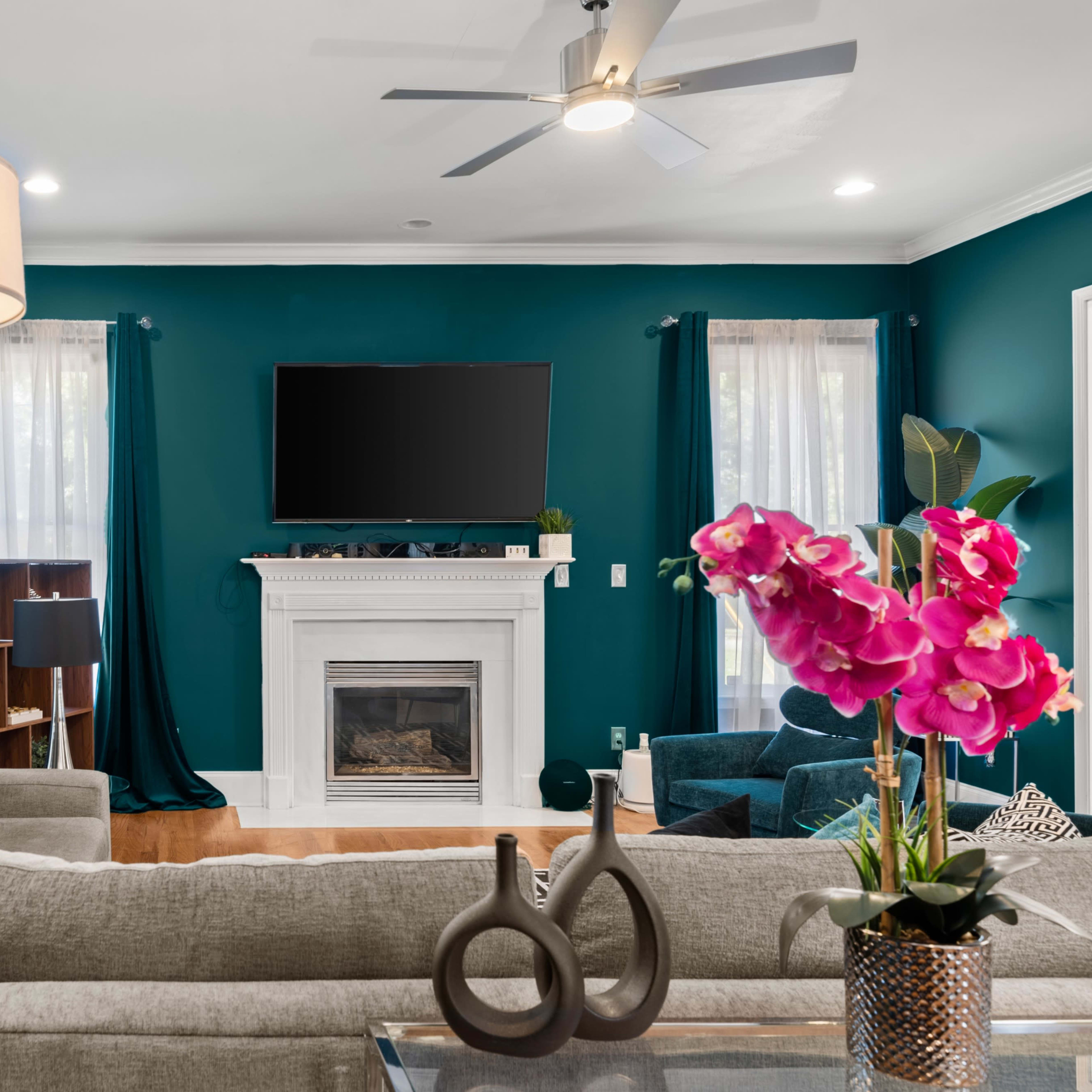 A modern living room features a grey sectional sofa, a fireplace with a TV above it, and teal curtains framing the windows.