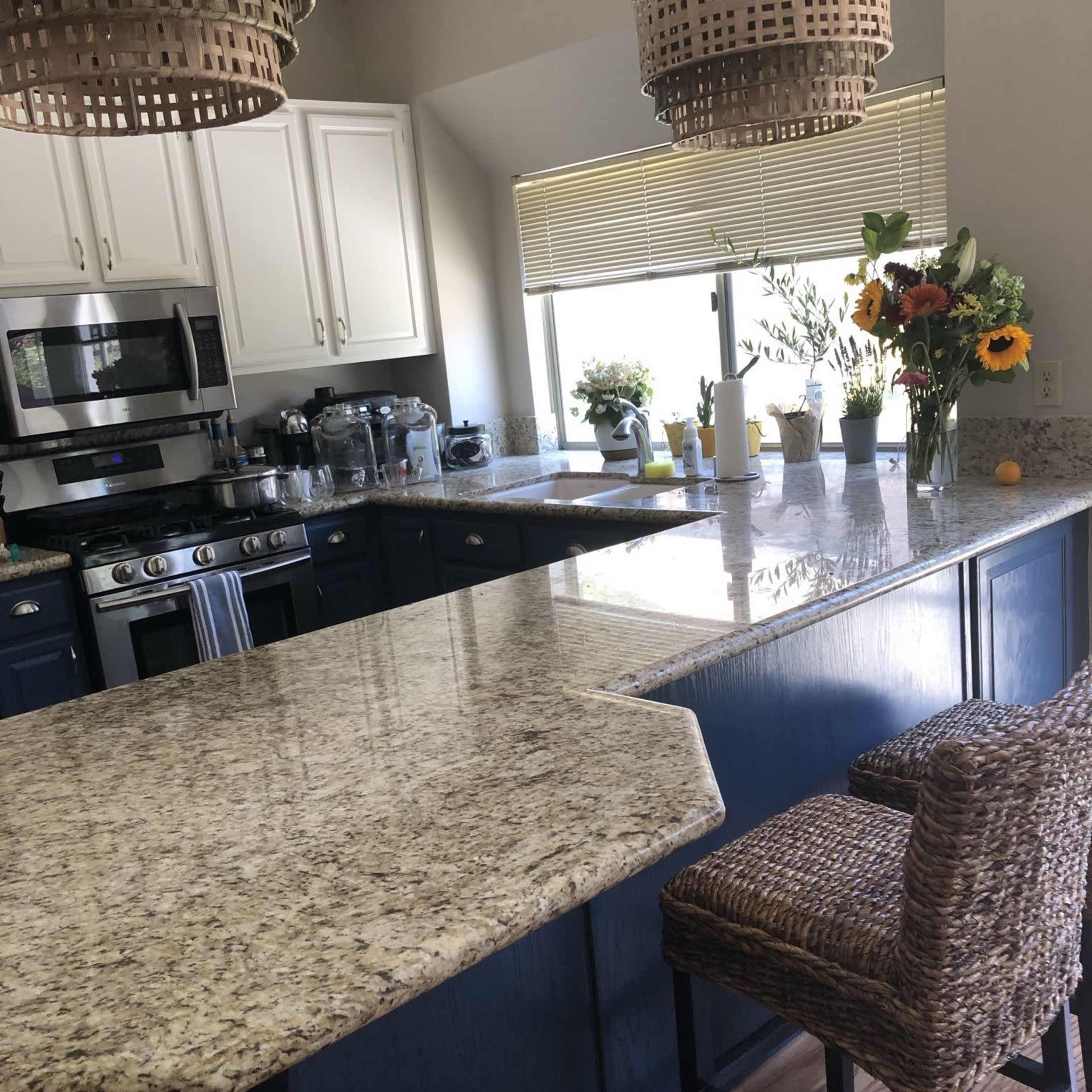 A kitchen with a large granite countertop, blue cabinets, two woven bar stools, and pendant lights hanging above.