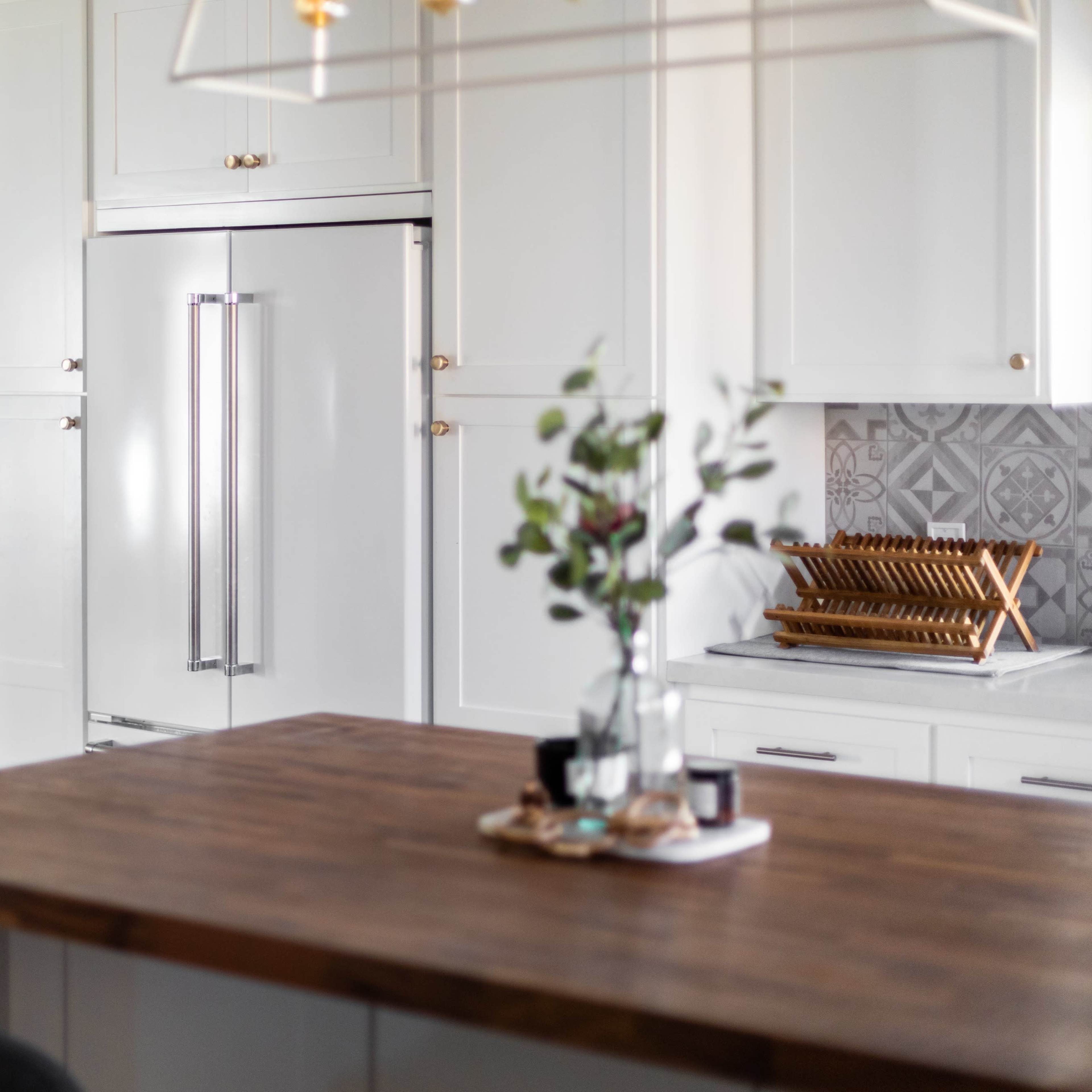 A modern kitchen features a large wooden island, a stainless steel refrigerator, and a vase with greenery on the counter, with a living area visible in the background.