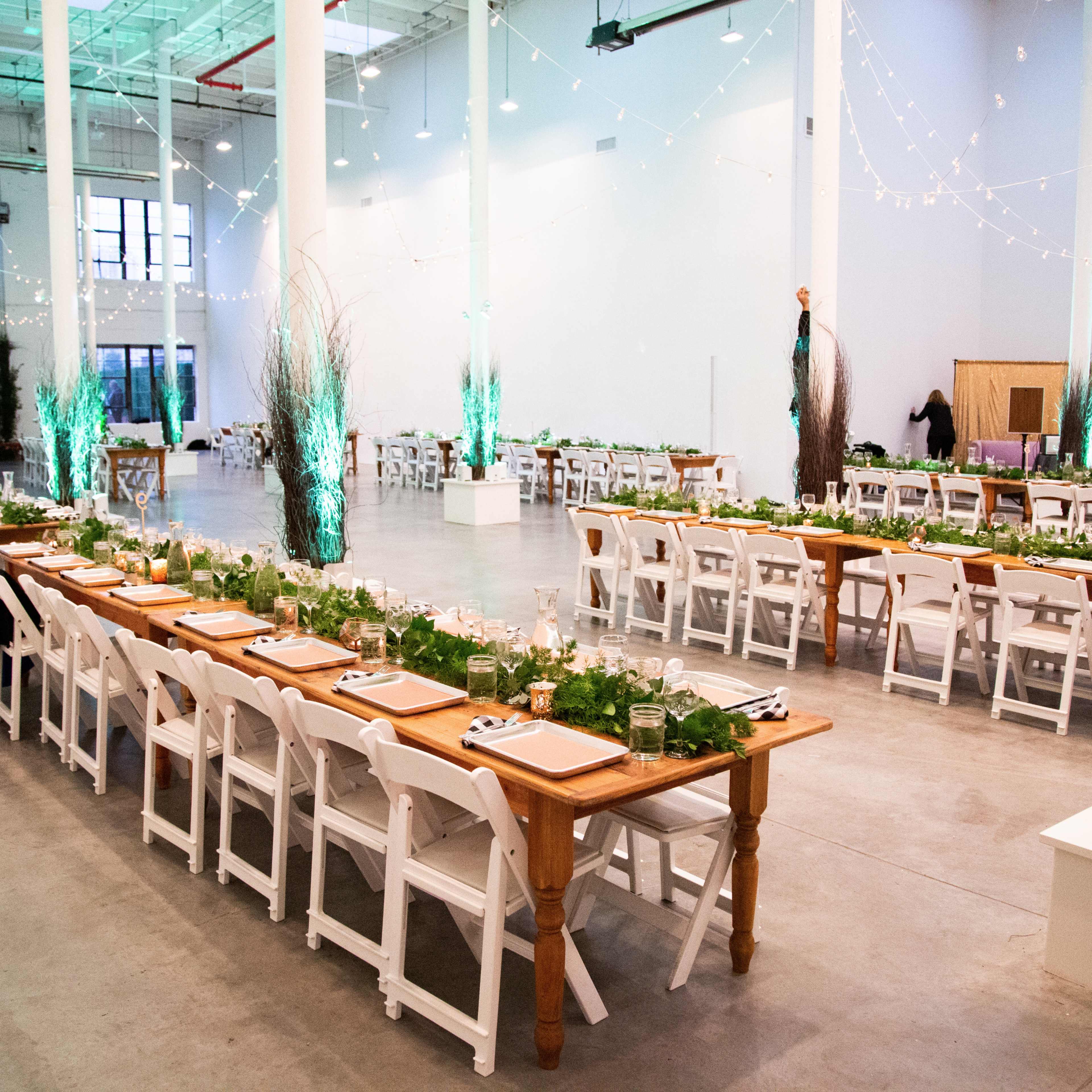 A spacious indoor venue features long tables set for a dinner event, adorned with greenery and illuminated by string lights.