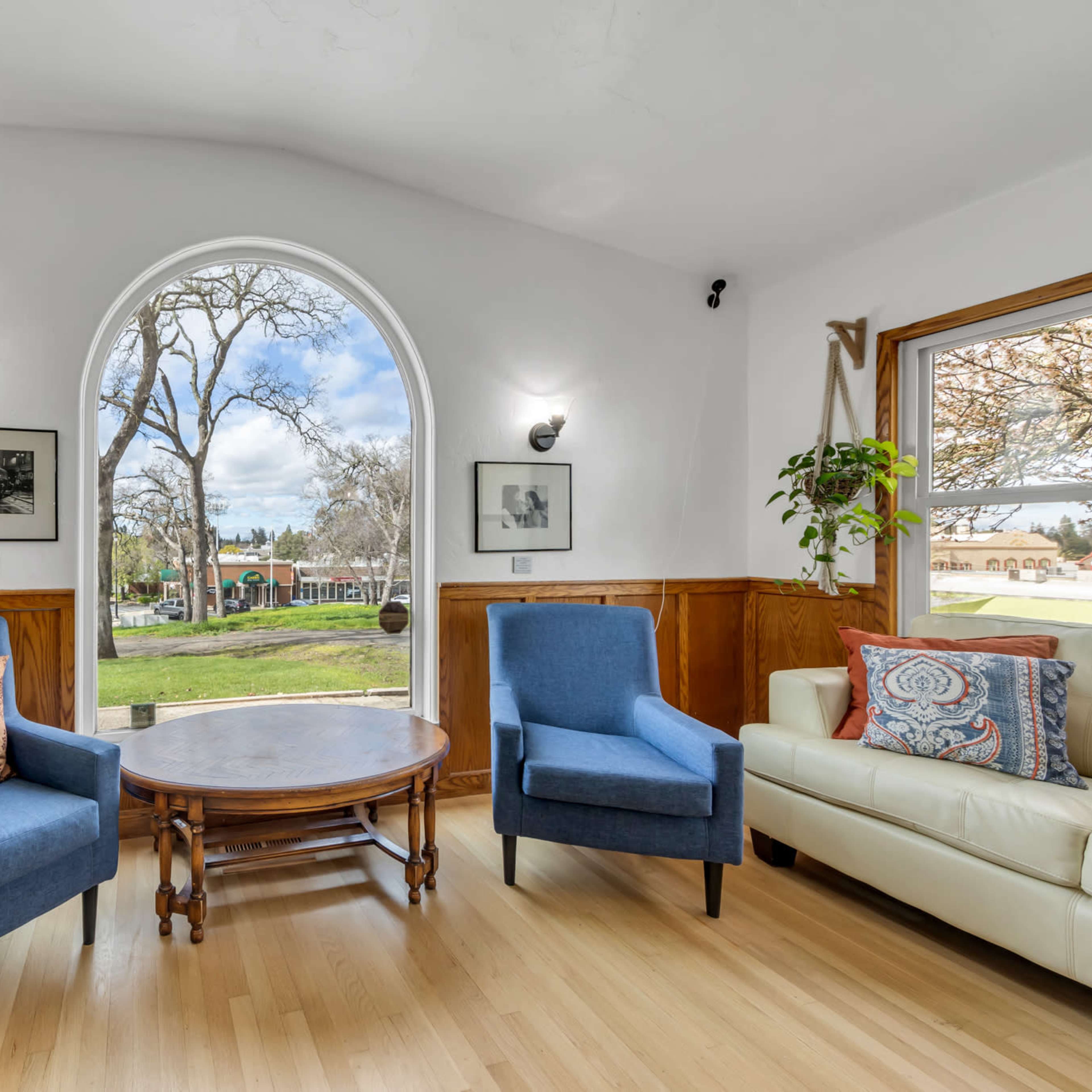 A cozy room with two blue chairs, a round wooden table, and a large window offering a view of a tree-lined outdoor area framed by wooden walls and black-and-white photographs.