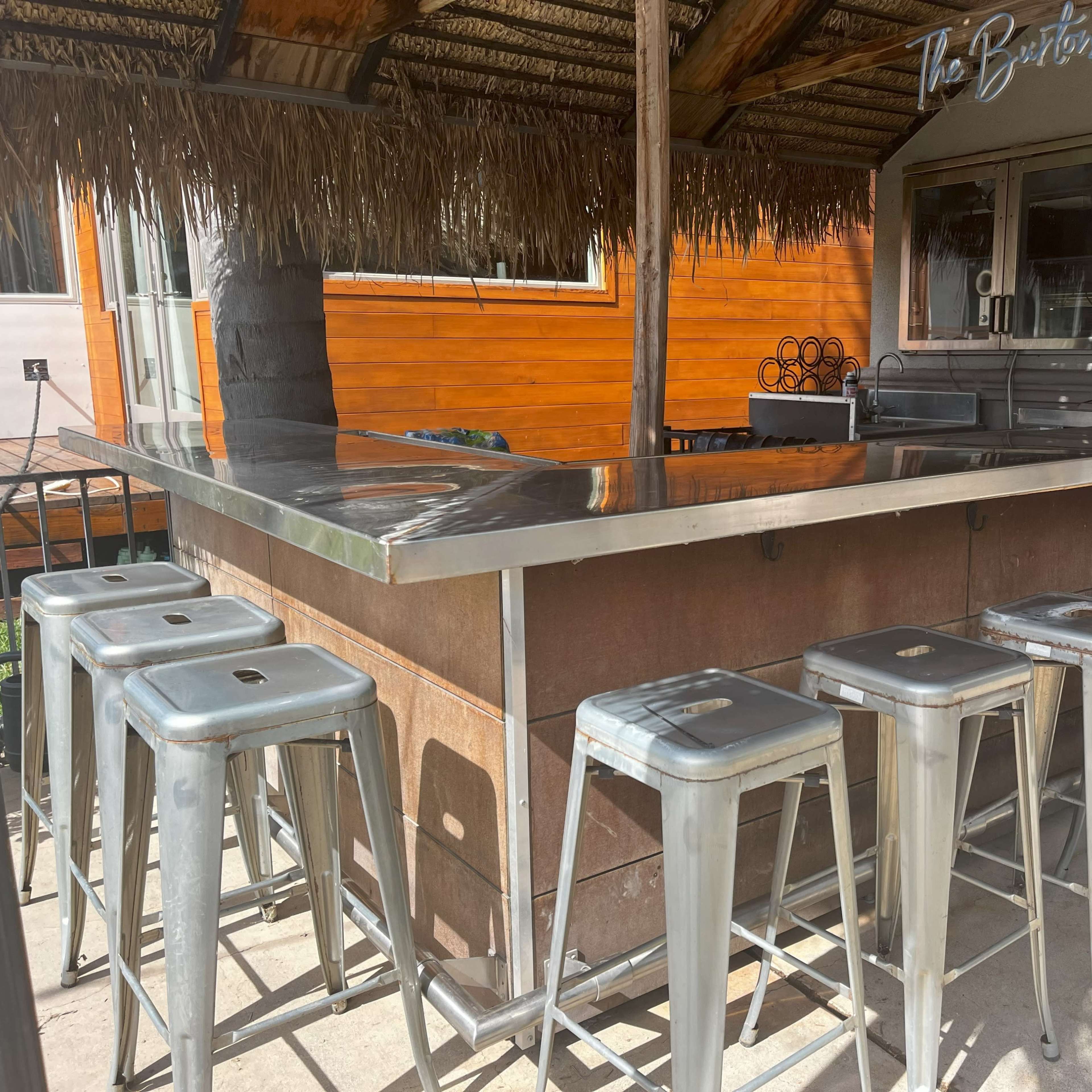 An outdoor bar with a metal countertop and several metal stools aligned in front.