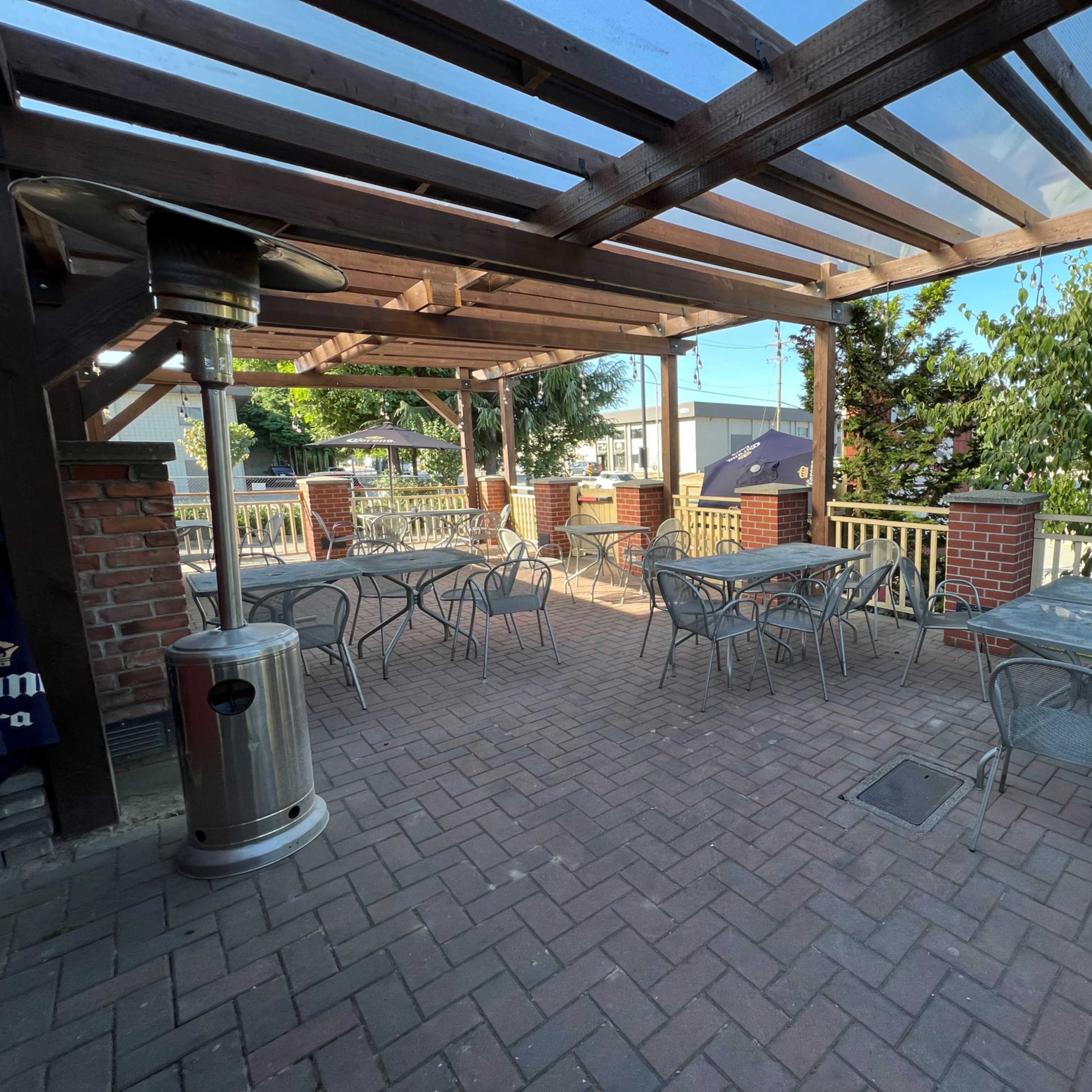 An outdoor patio area with metal tables and chairs, sheltered by a wooden pergola with a glass roof and brick columns.