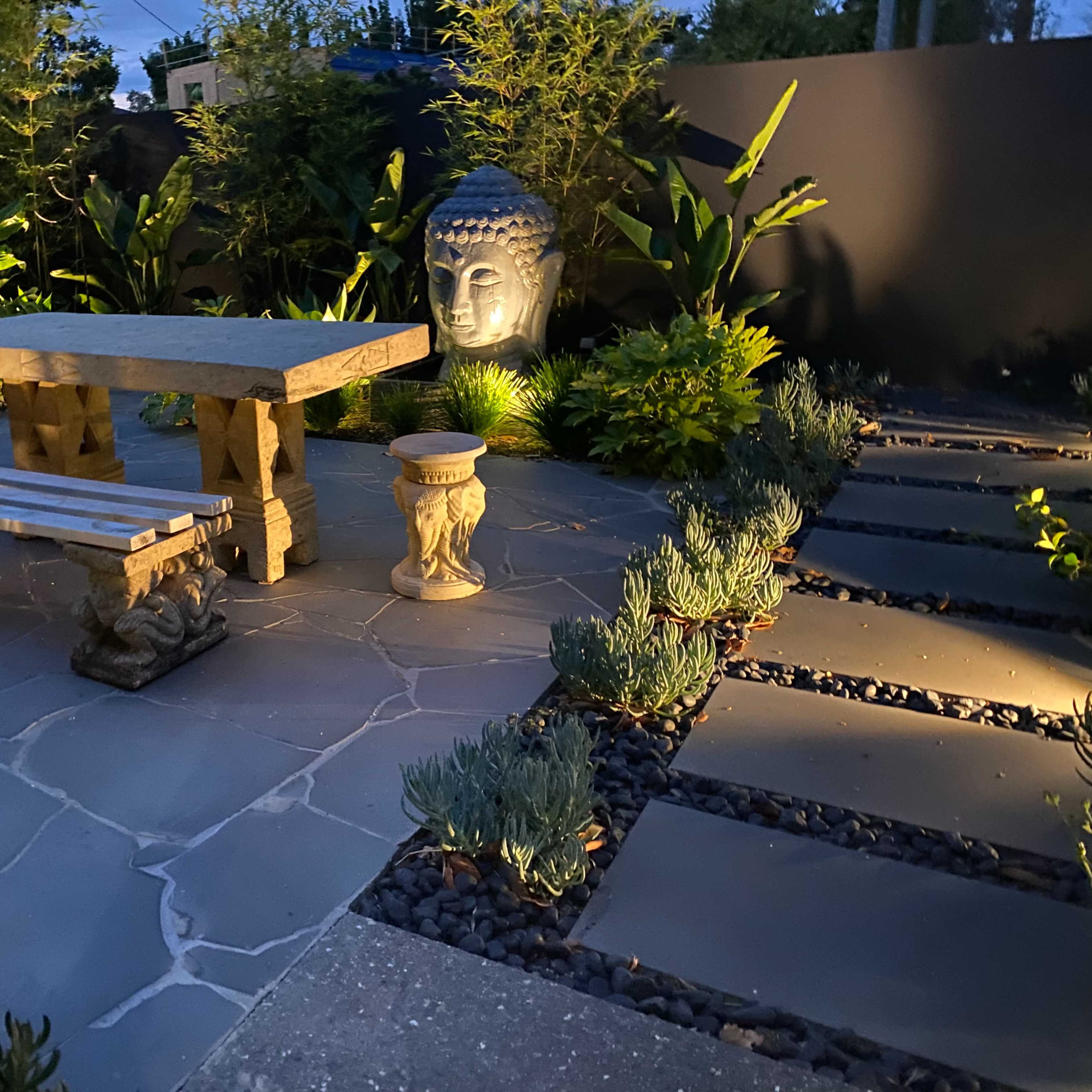 A garden scene featuring a stone table and benches surrounded by lush greenery and a statue of Buddha, illuminated by soft lighting.