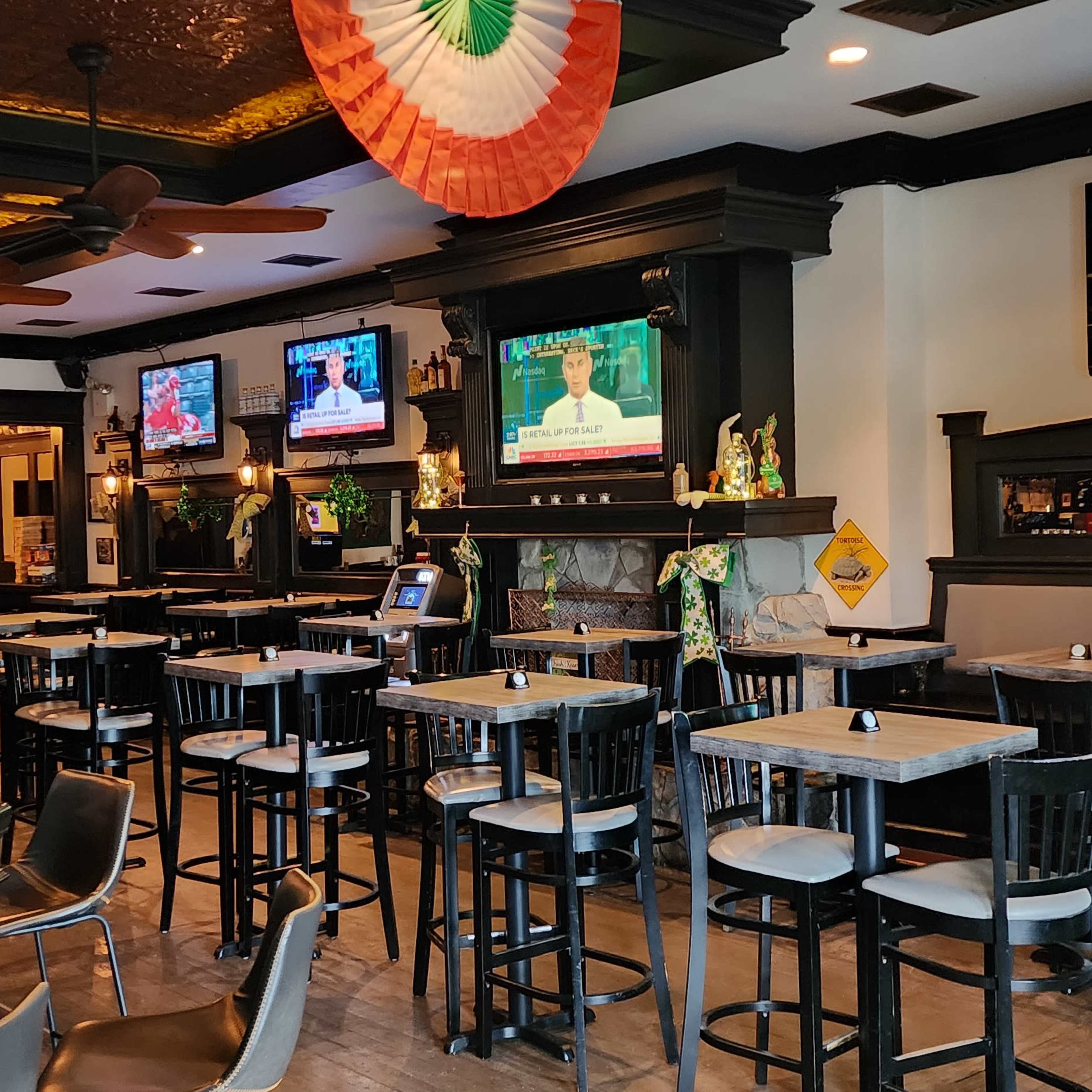 An Irish pub interior with multiple television sets displaying news, surrounded by high tables and bar stools under festive decorations.