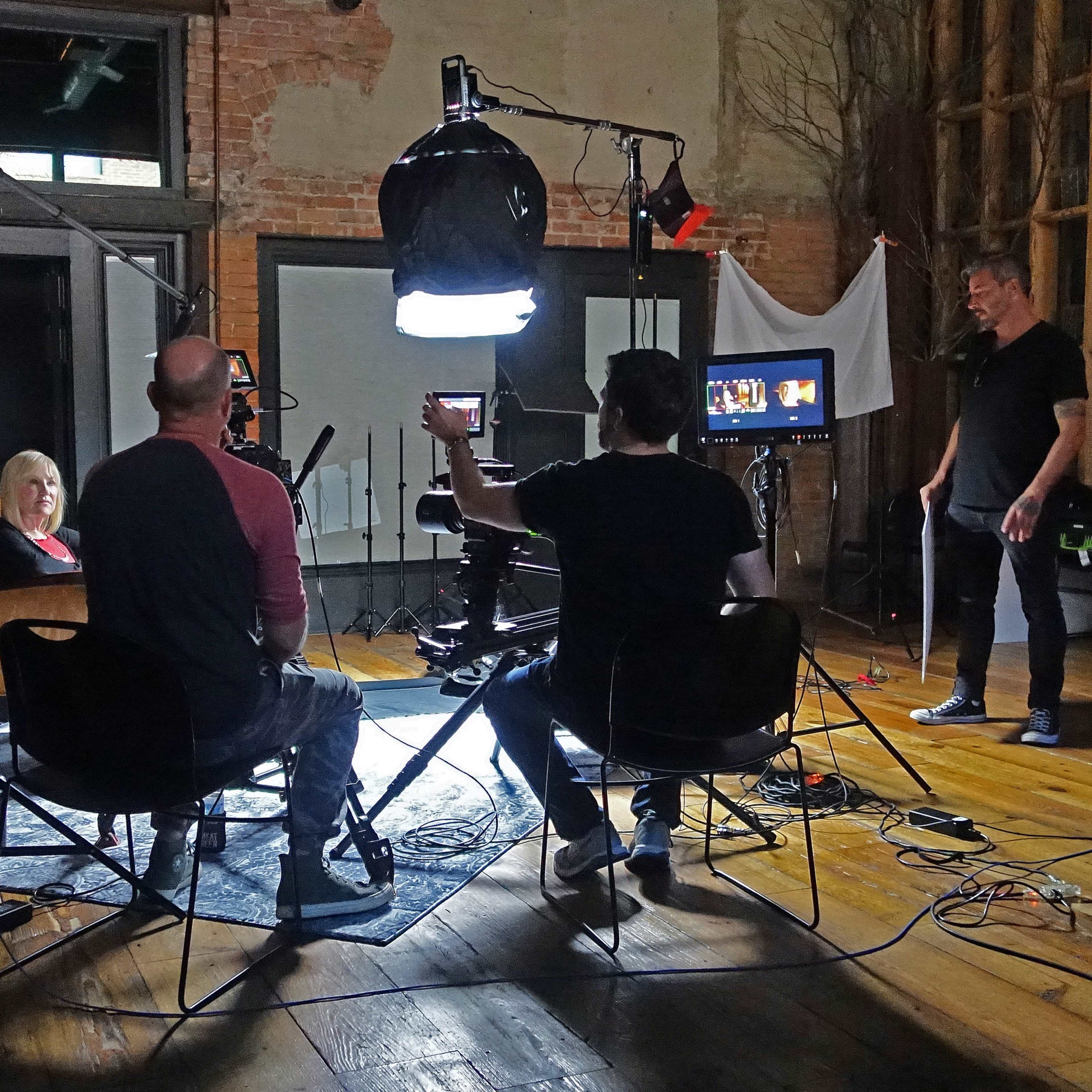 A film crew operates in a spacious studio, setting up cameras and lighting around a seated subject.