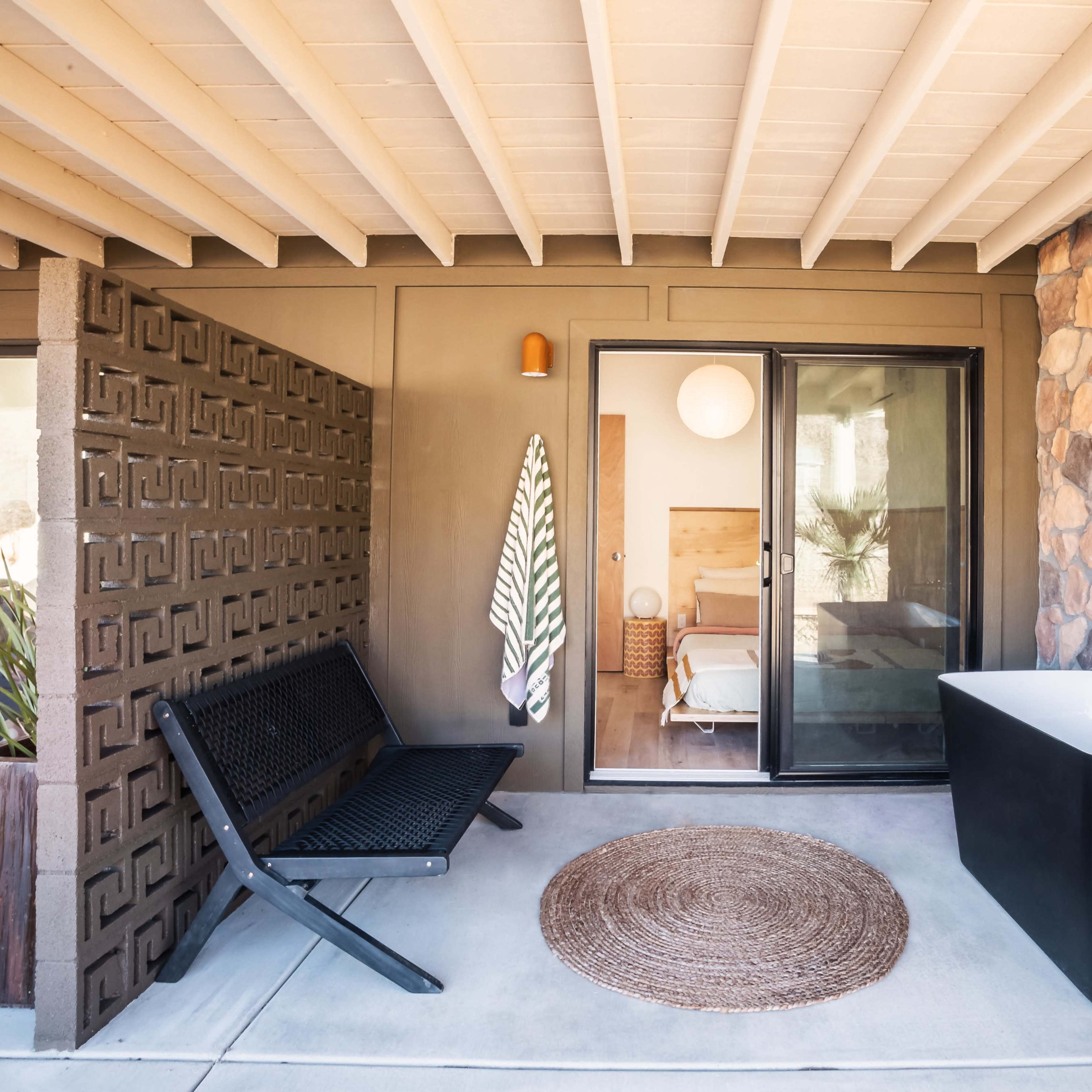 A cozy outdoor area featuring a black freestanding bathtub, a woven rug, a modern bench, and a wall made of stone and decorative blocks, leading into a bedroom.
