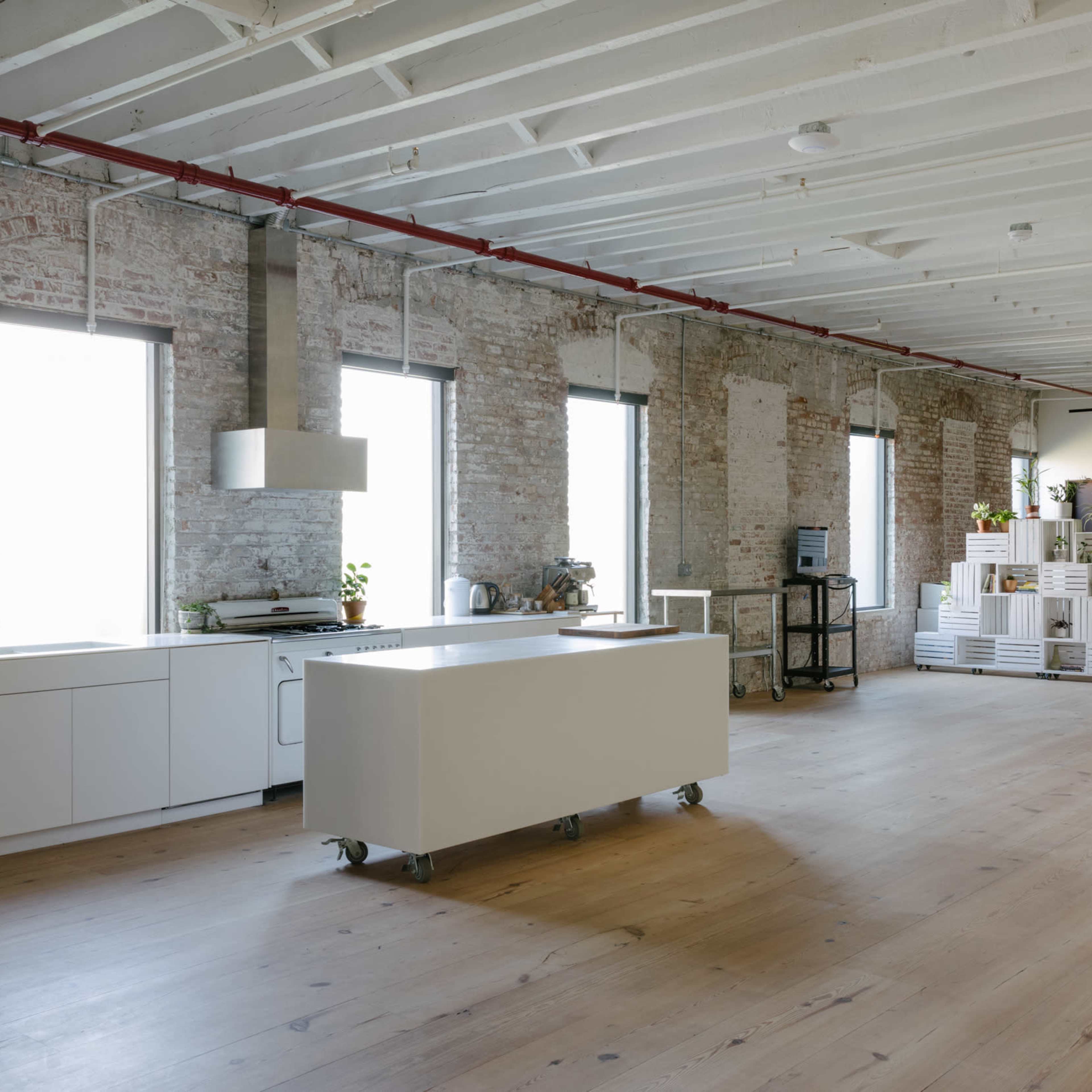 A spacious industrial-style kitchen area with white brick walls, large windows, and a central island on wheels.