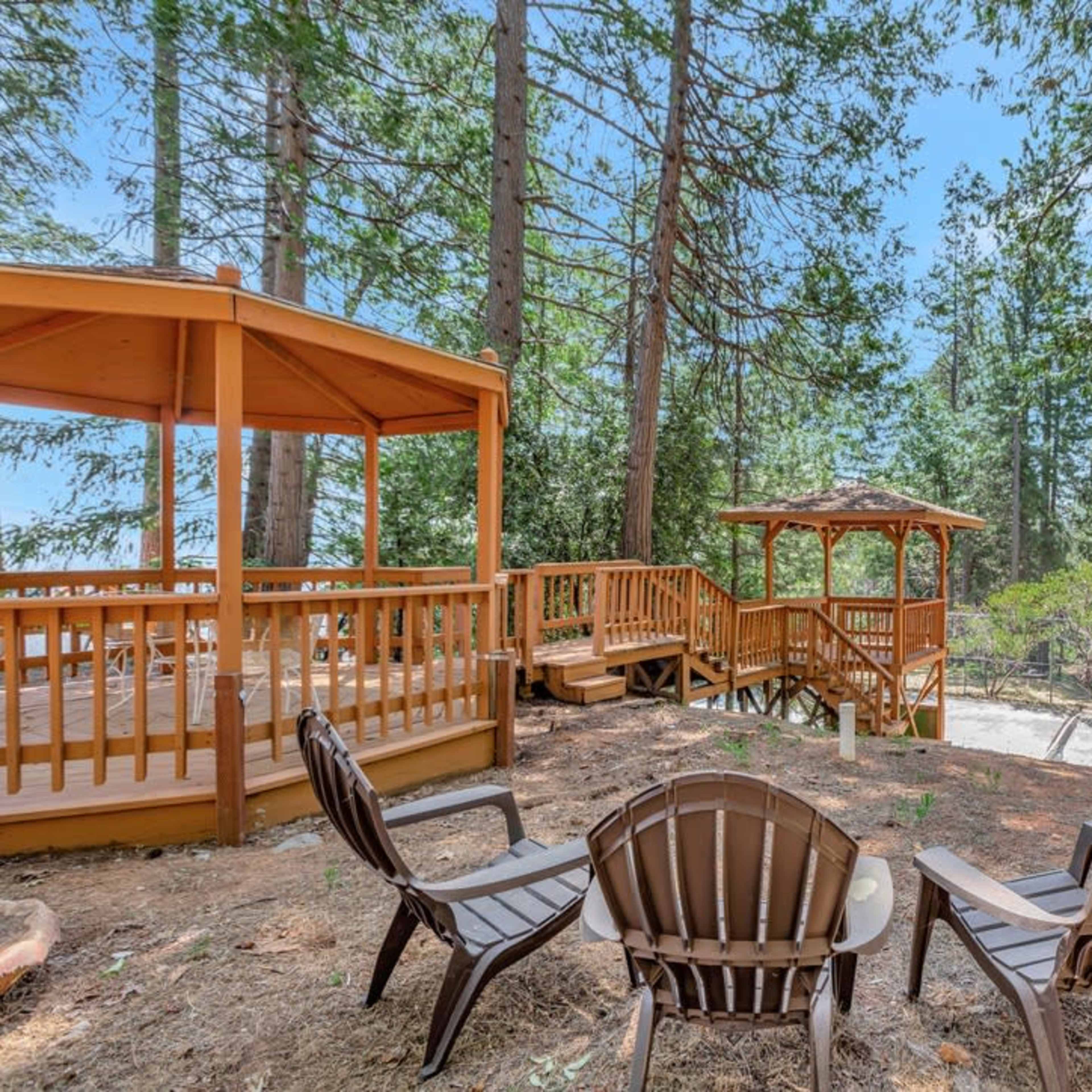 A wooden gazebo and deck area are surrounded by tall pine trees in a forested setting.