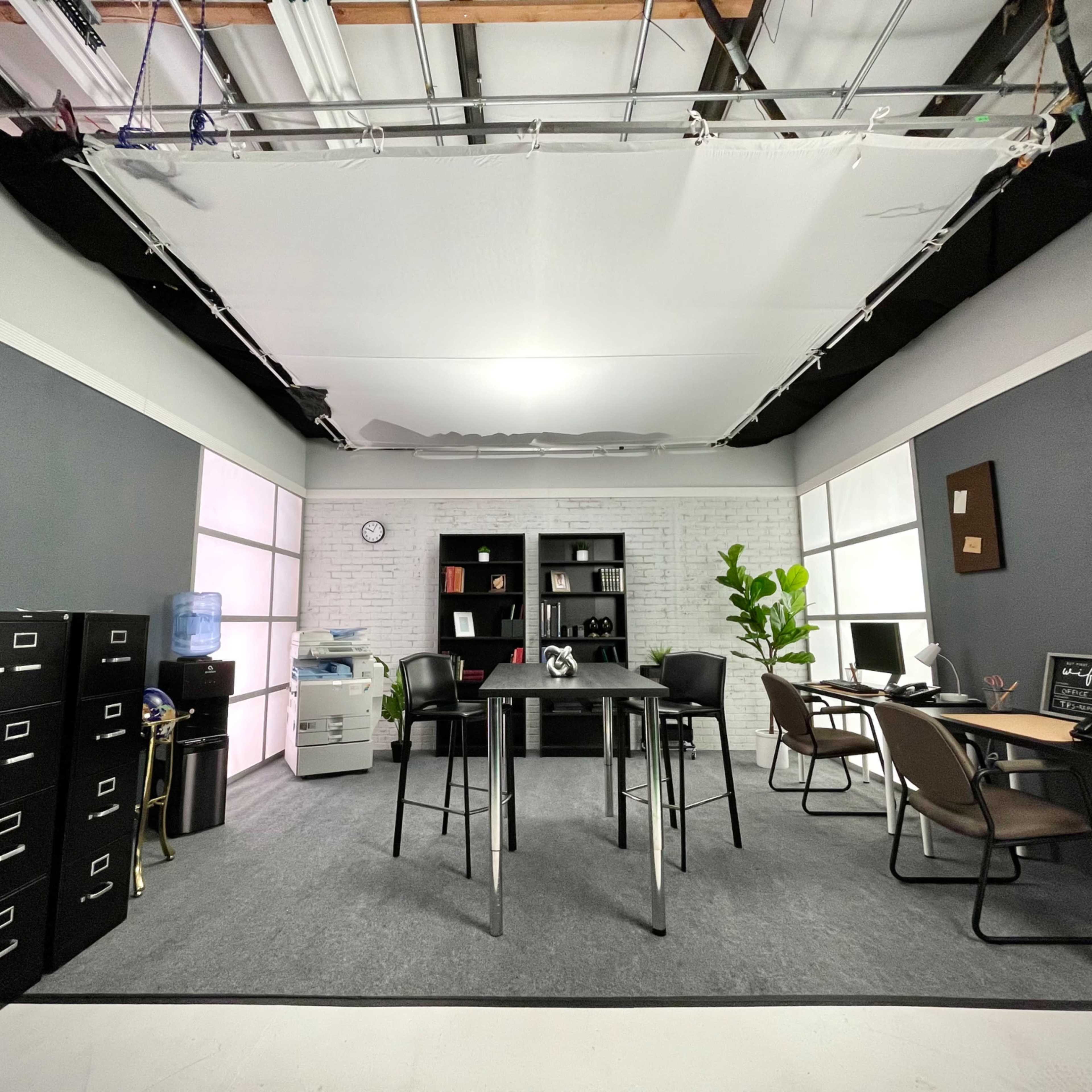 A modern office space with a central table, filing cabinets, chairs, and a desk setup against a backdrop of gray walls and large windows.