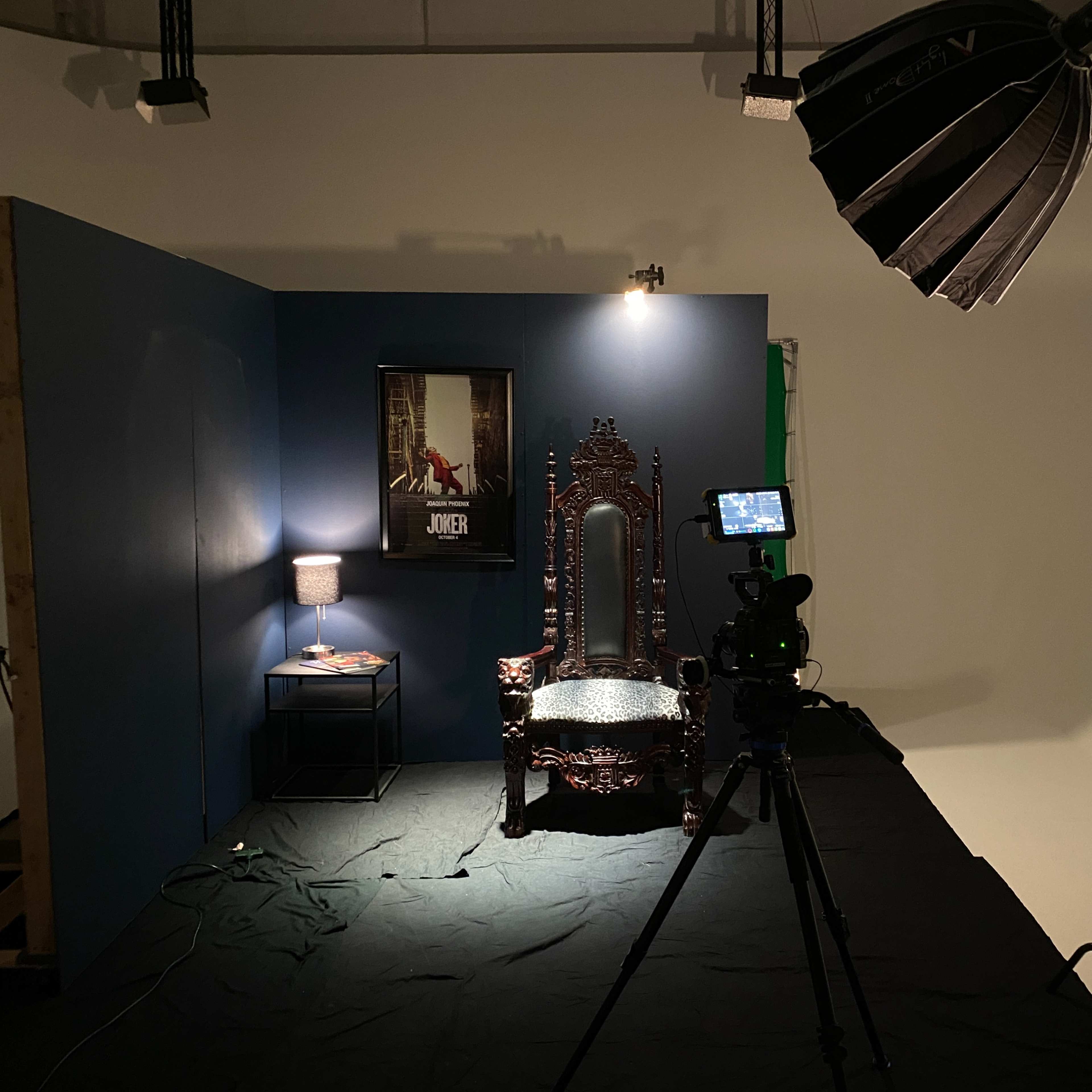 A photography studio setup with a decorative throne-like chair, a table lamp, a poster on the wall, and a camera on a tripod pointed at the chair.