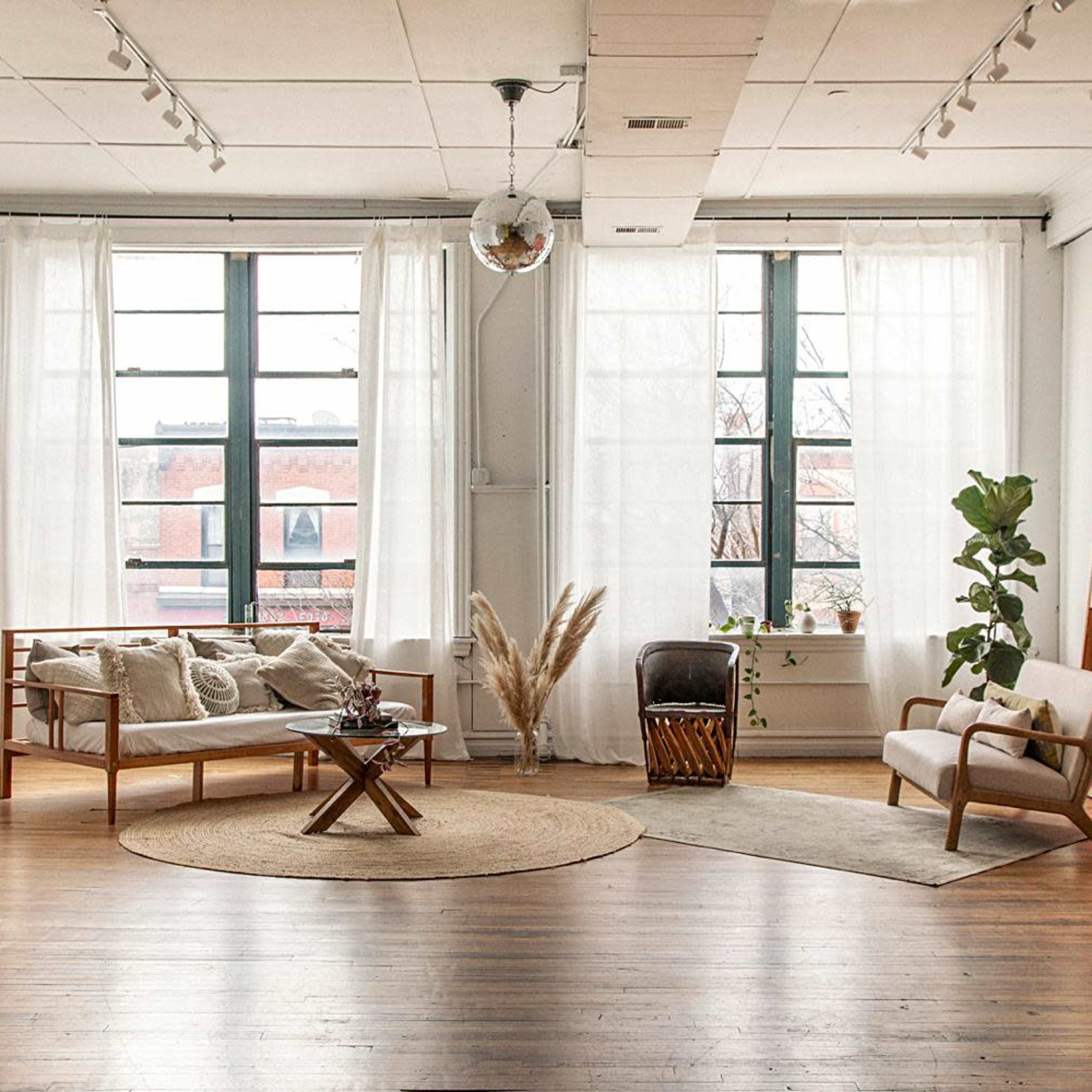 A bright, spacious living room with large windows, wooden floors, a cozy seating area, and decorative plants.