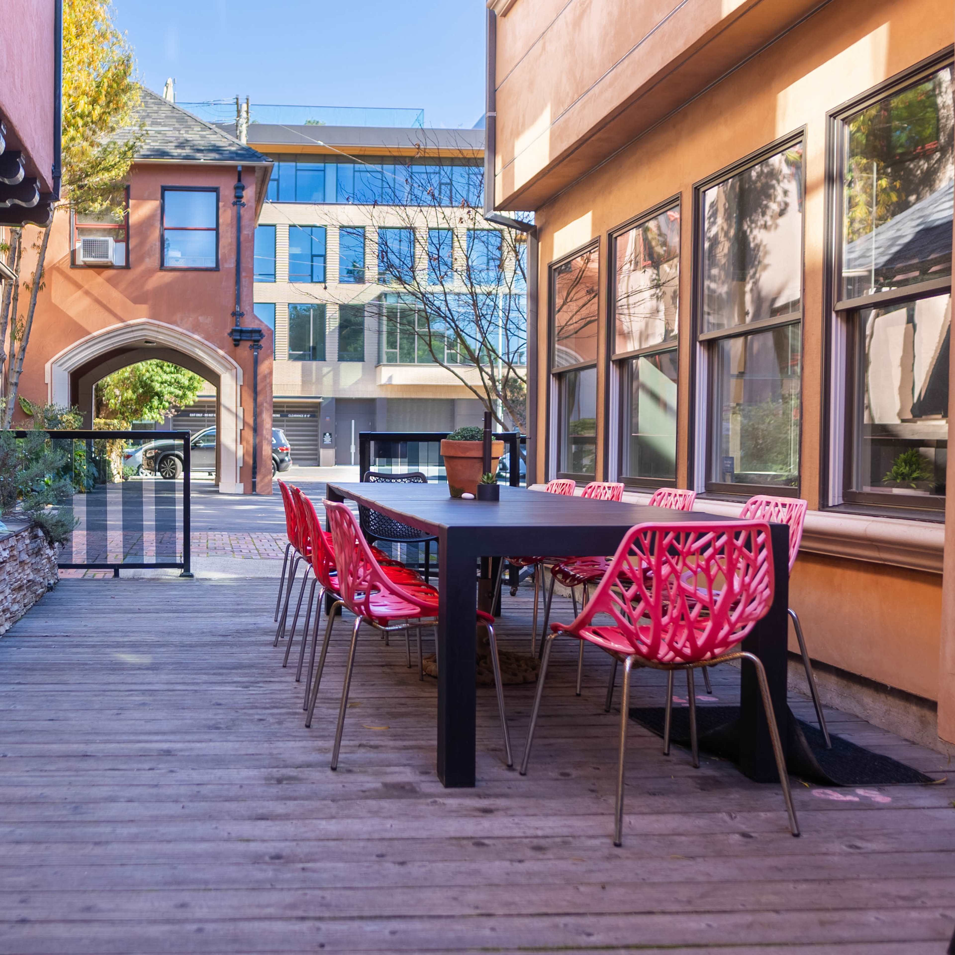 A wooden patio features a black table surrounded by red chairs, with buildings visible in the background.