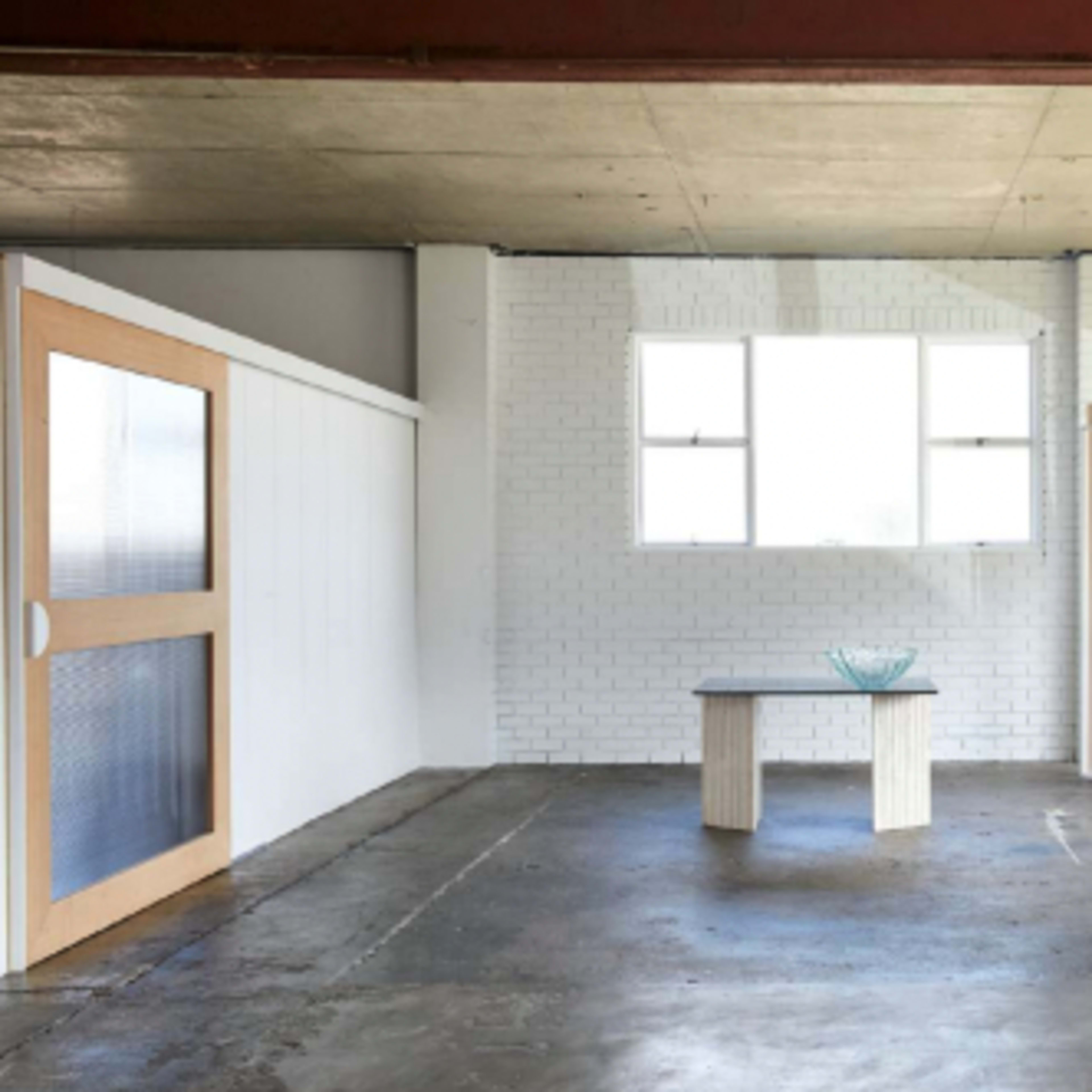 An empty room features a small table in the center, with large windows allowing natural light to enter.