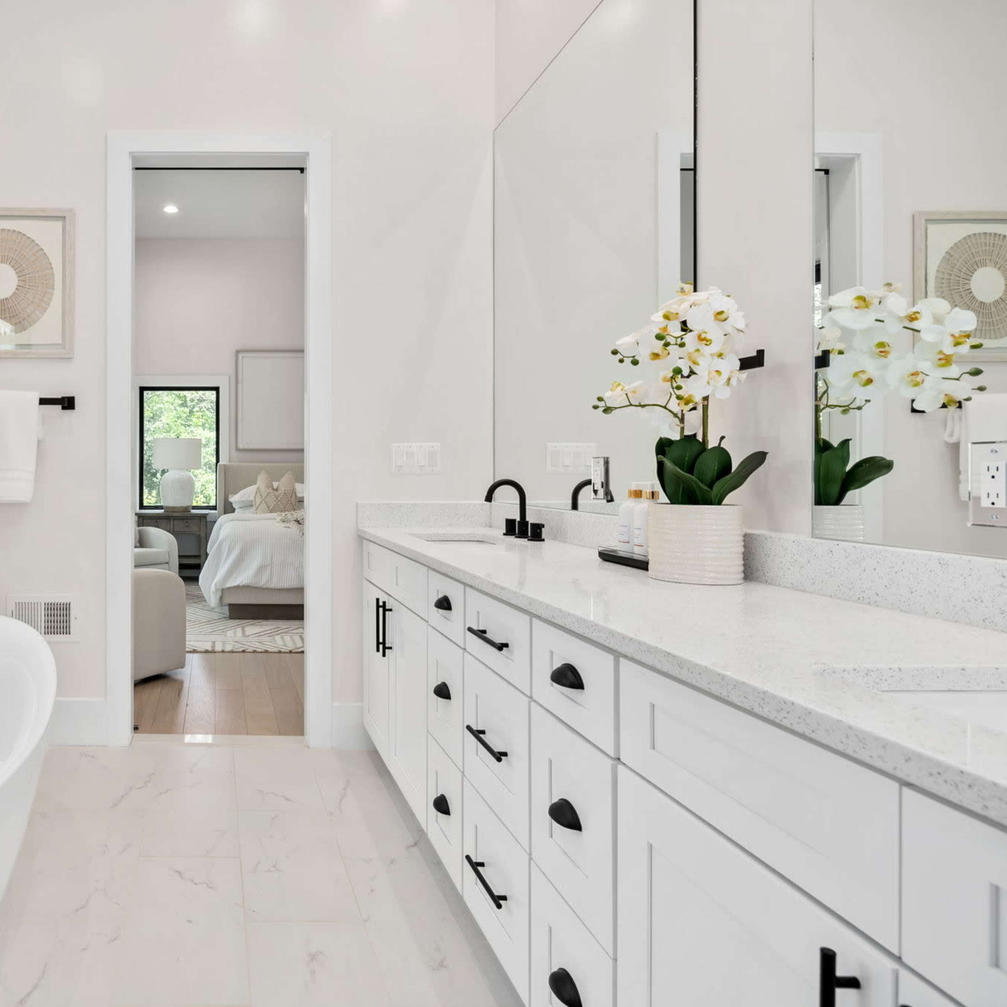A modern bathroom with a freestanding bathtub, a dual sink vanity with black fixtures, and a large mirror, all set against light-colored walls and a view of a bedroom through an open door.