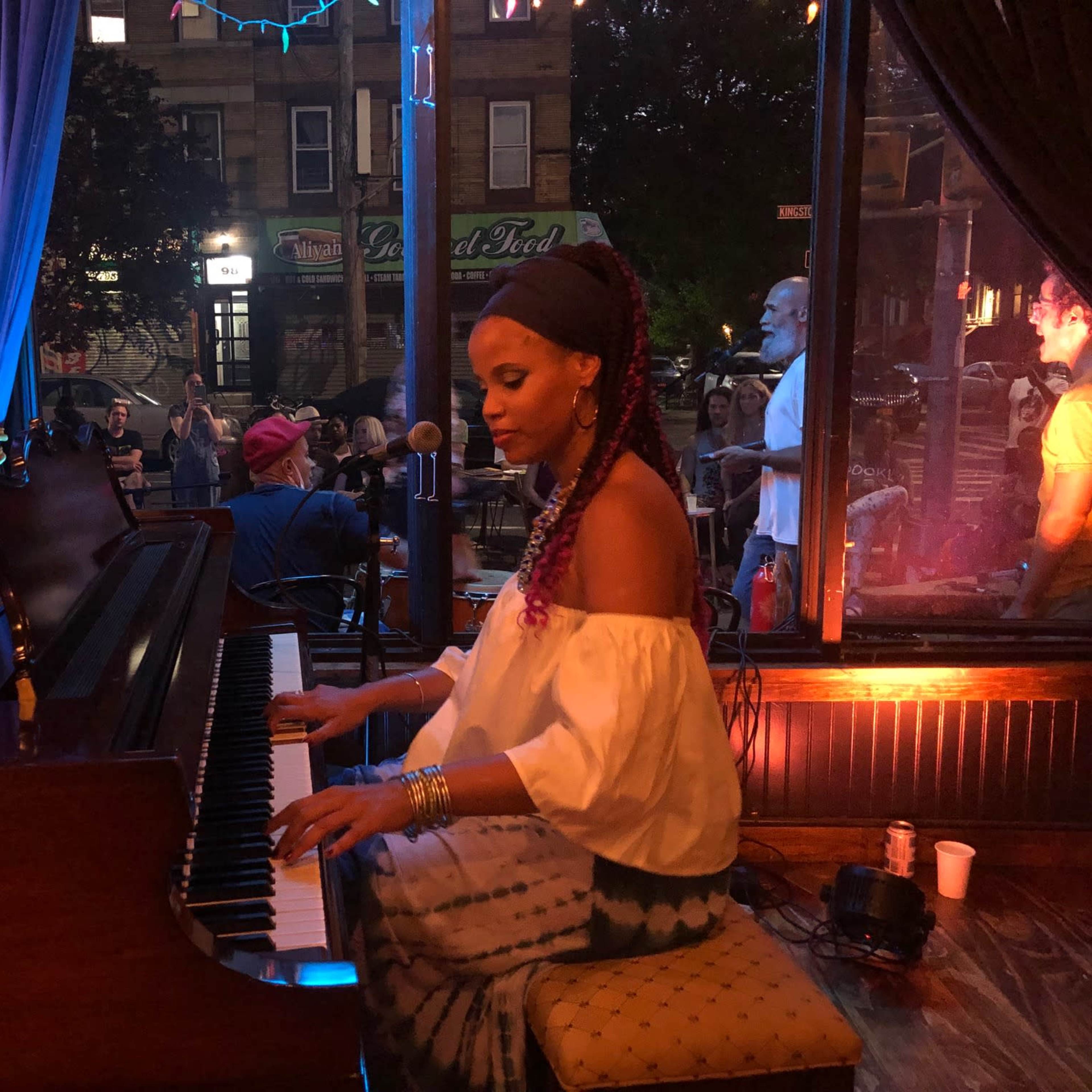 A musician plays the piano in a dimly lit venue with patrons visible in the background.