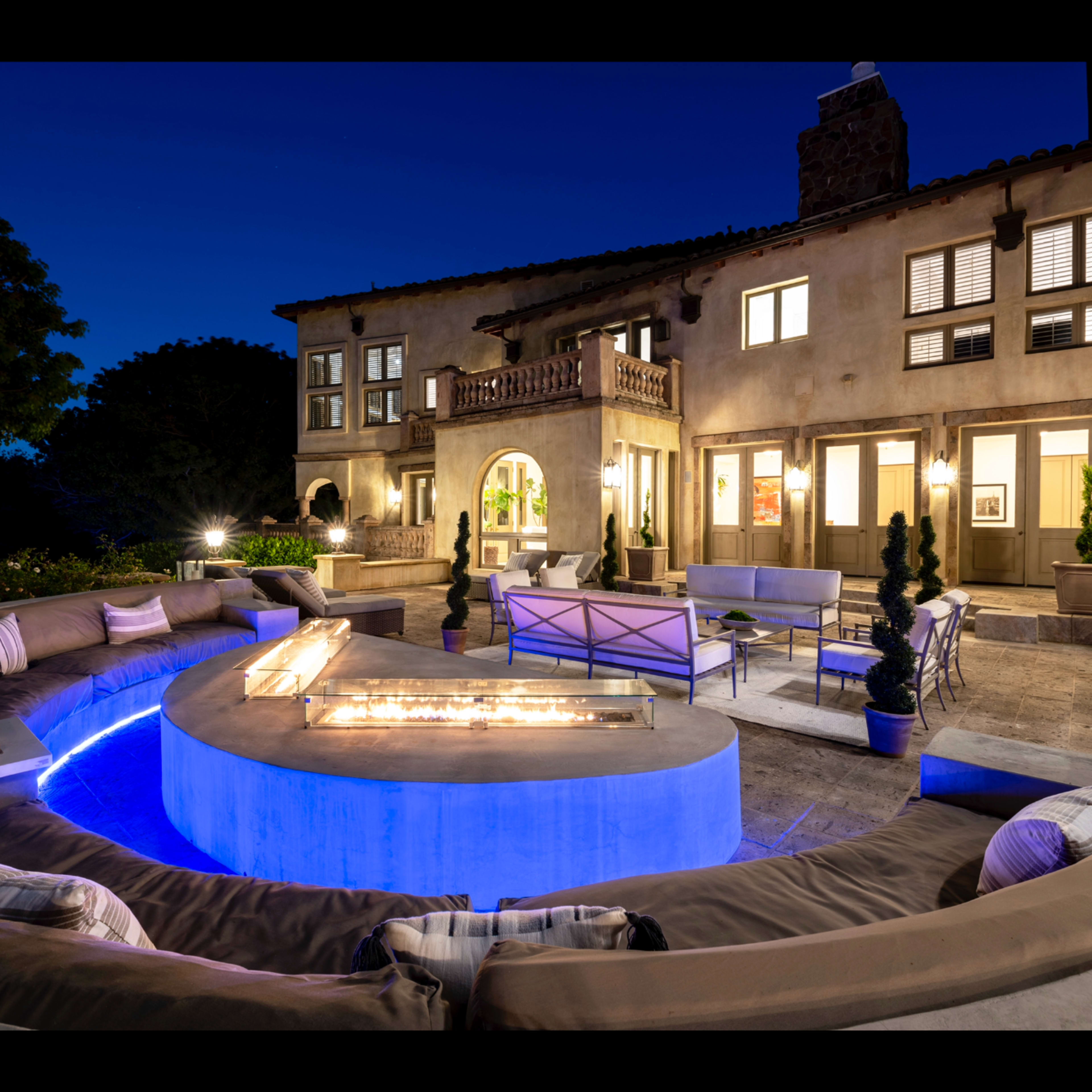 A modern outdoor living space with a circular fire pit surrounded by a curved bench, set against a backdrop of a well-lit multi-story home at dusk.