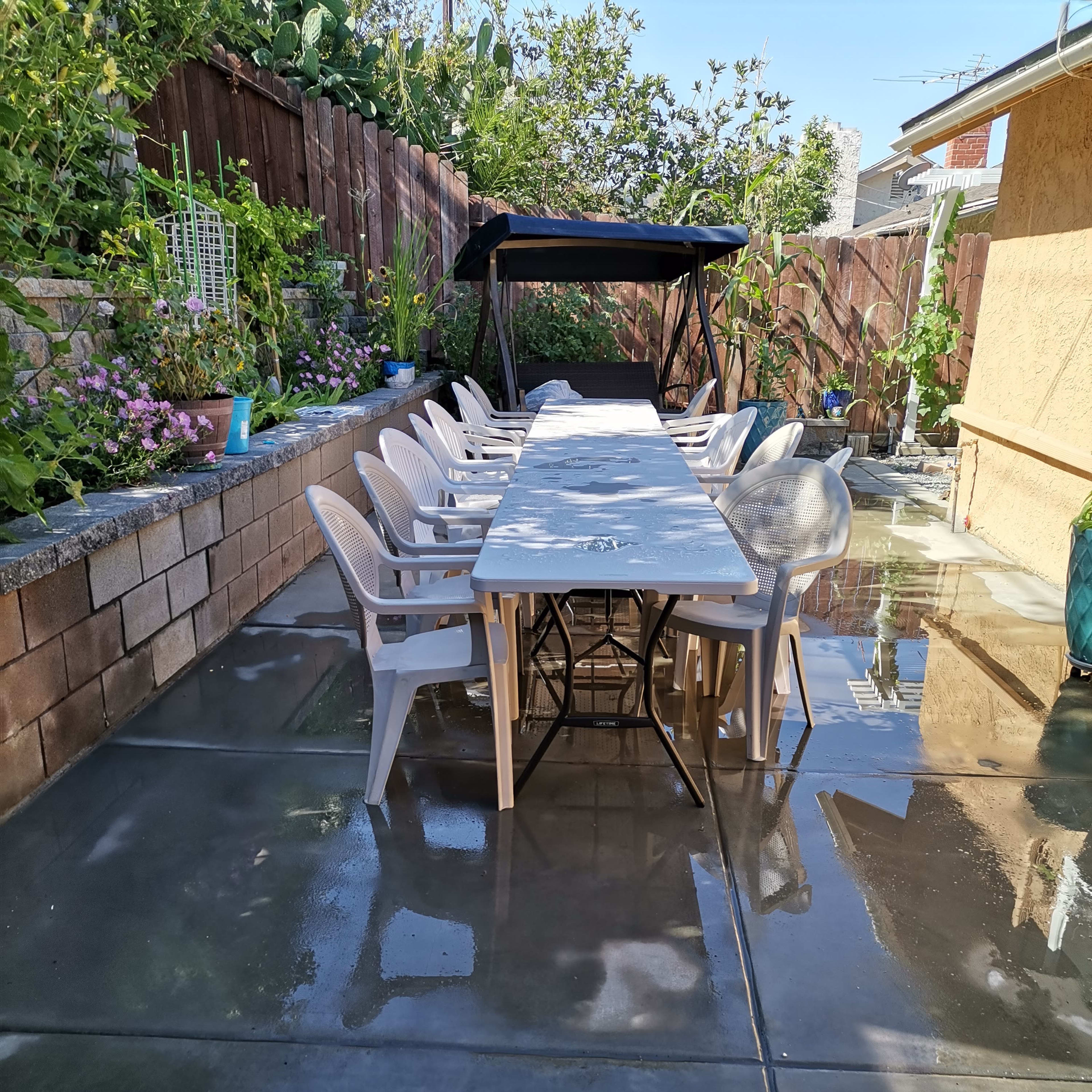 A long table with chairs is set up on a wet concrete patio surrounded by greenery and a shaded seating area.