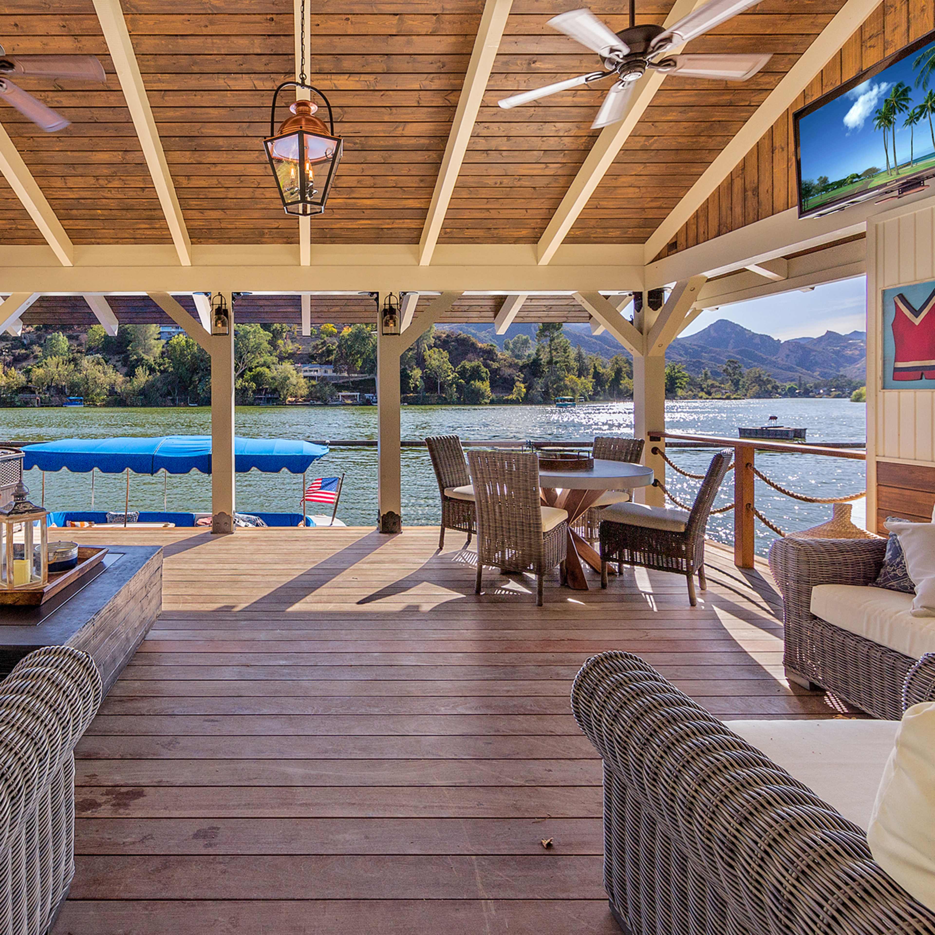 A spacious lakeside patio features a seating area with white sofas, a wooden coffee table, and a dining set, all overlooking the water and mountains.