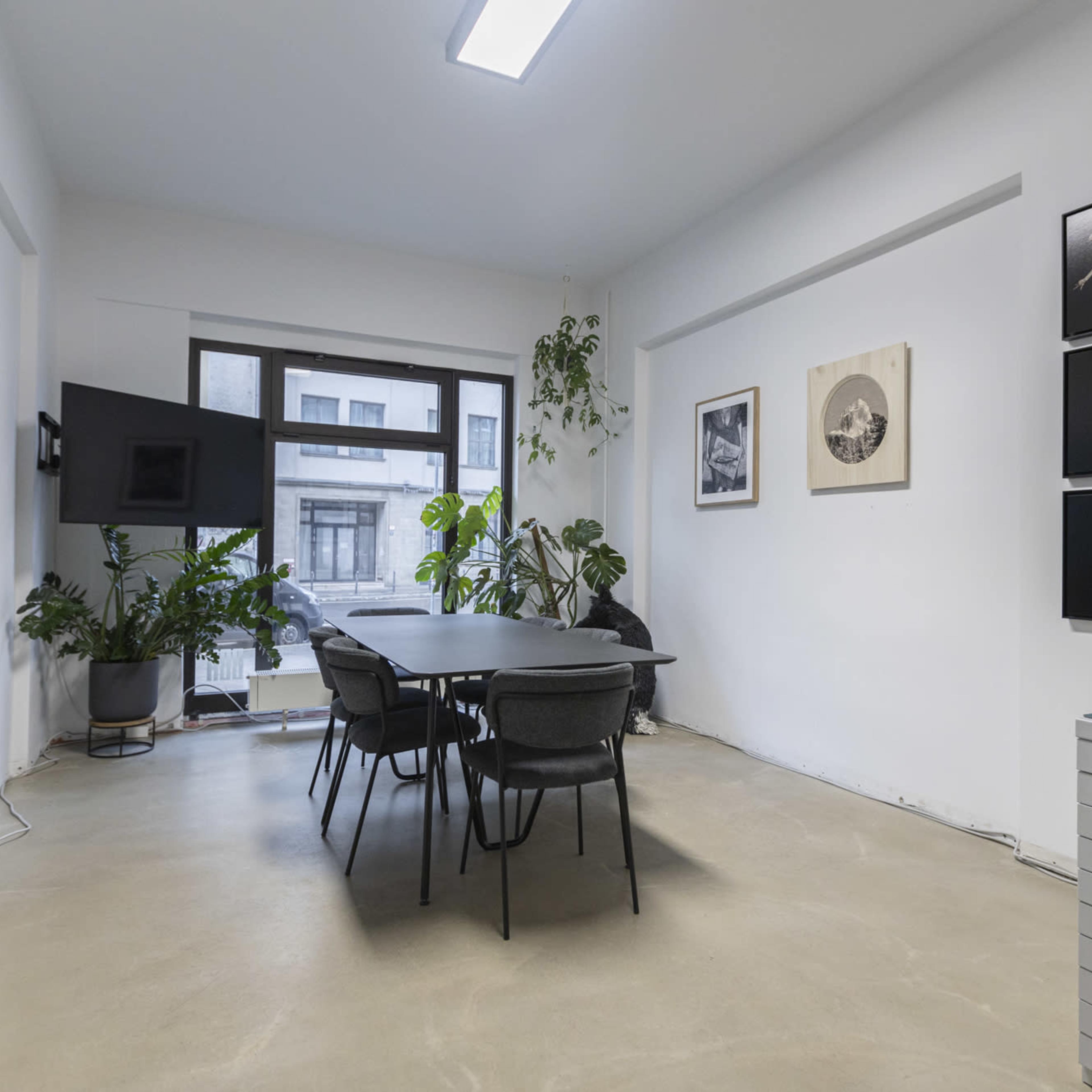 A well-lit meeting room featuring a large table surrounded by black chairs, with various artworks hanging on the walls and indoor plants adding greenery to the space.