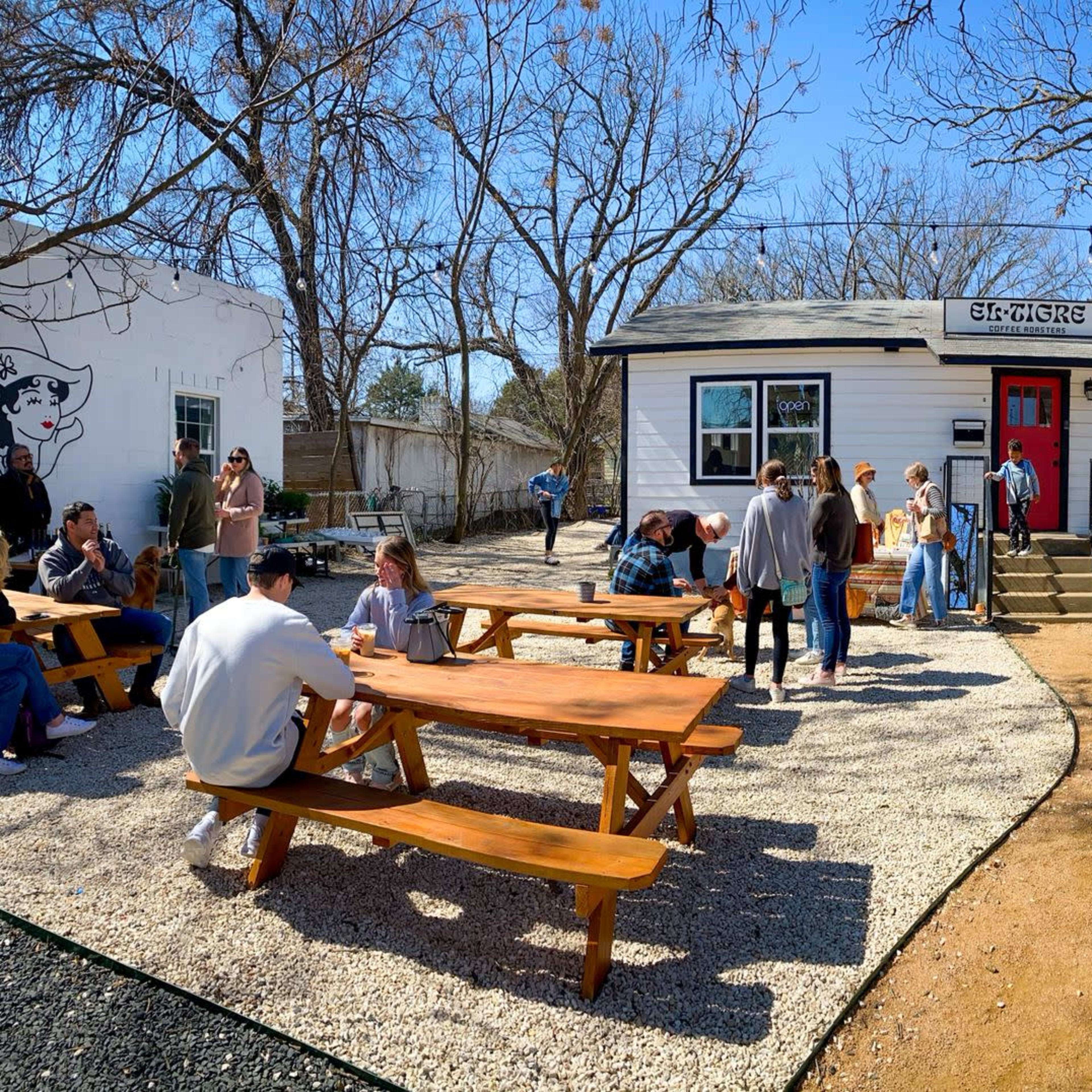 A group of people congregates outside a white building with a red door, surrounded by picnic tables and bare trees on a sunny day.
