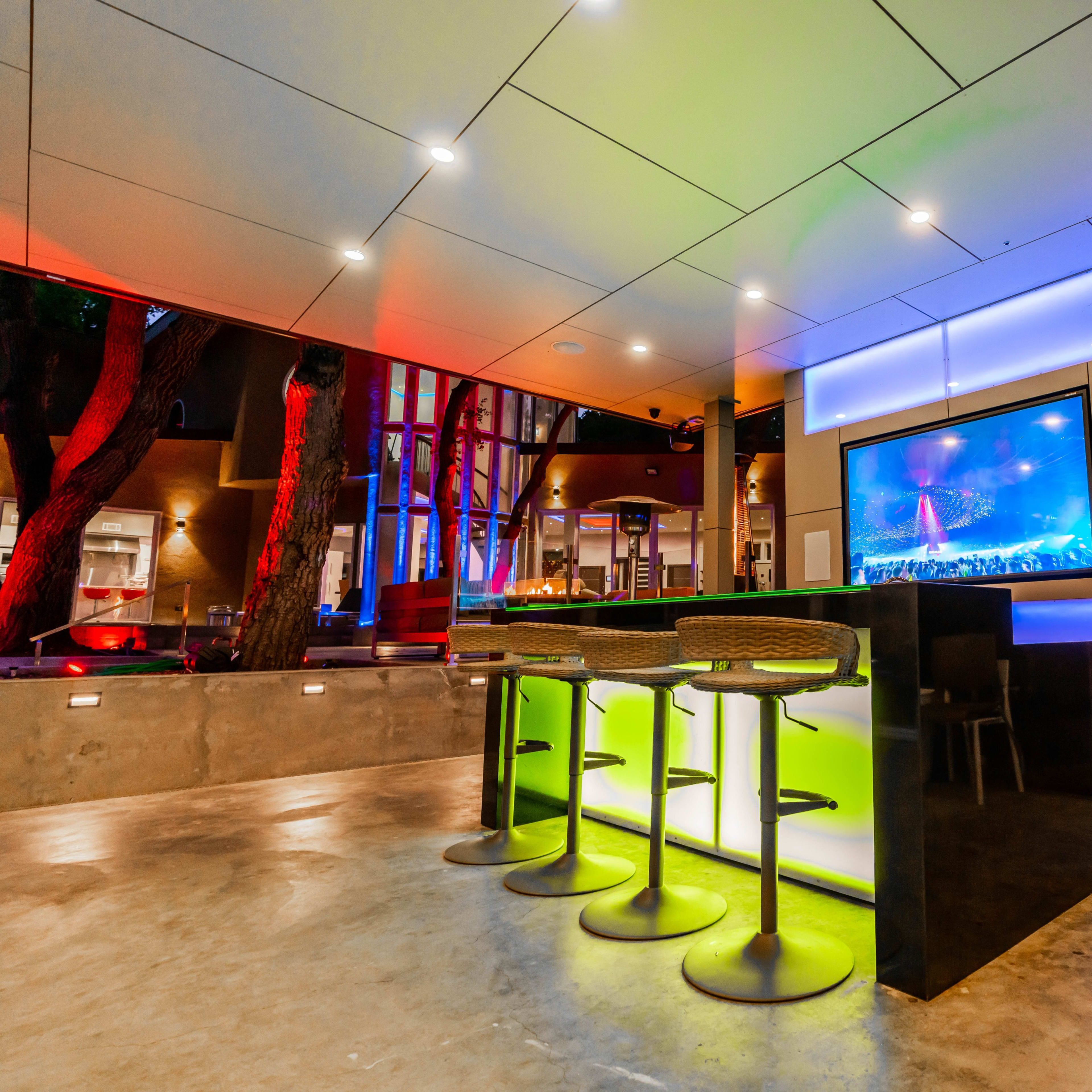 A modern bar area features illuminated stools and a large screen displaying a performance, surrounded by trees and a contemporary interior design.