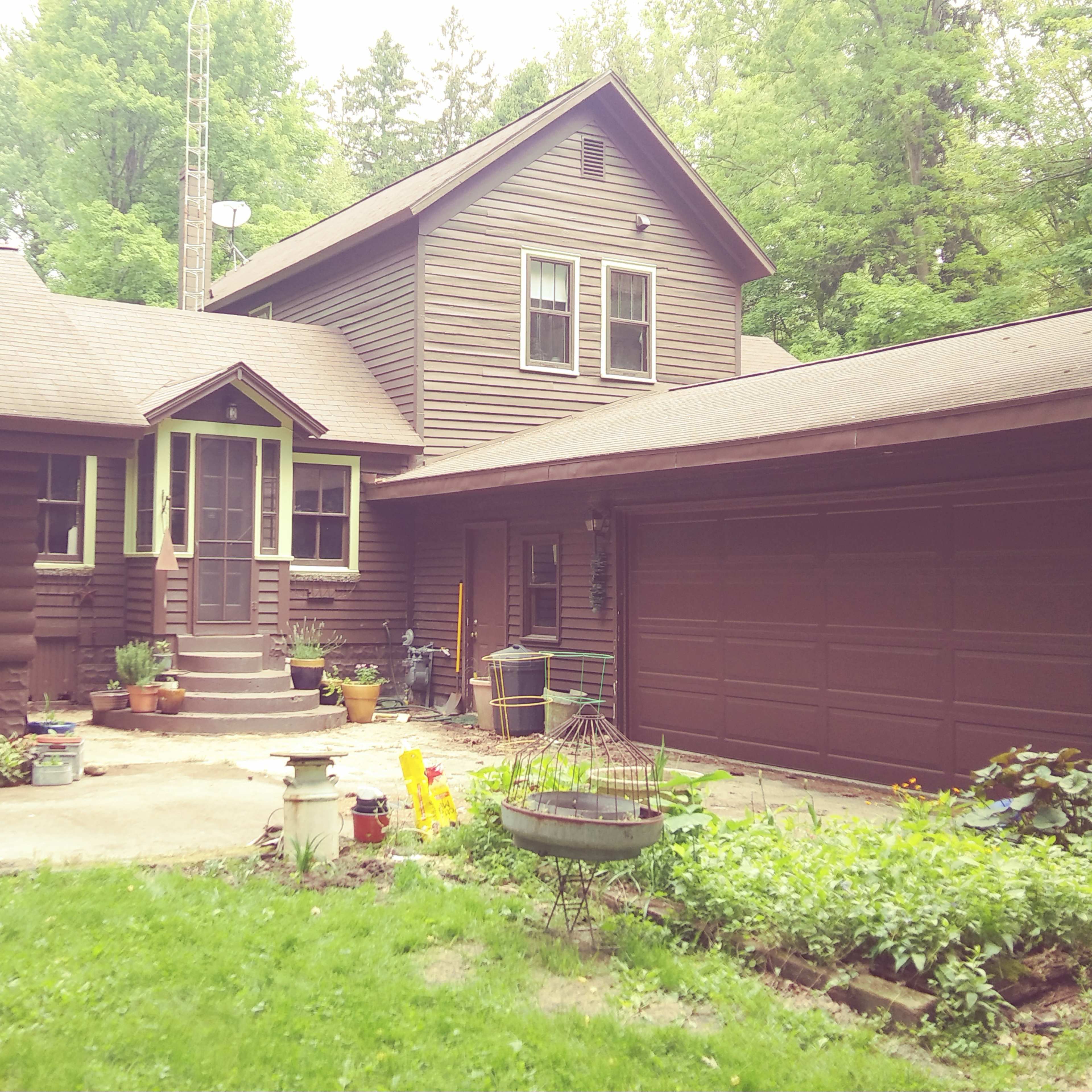 A two-story brown wooden house with a porch, a garage, and a landscaped yard.