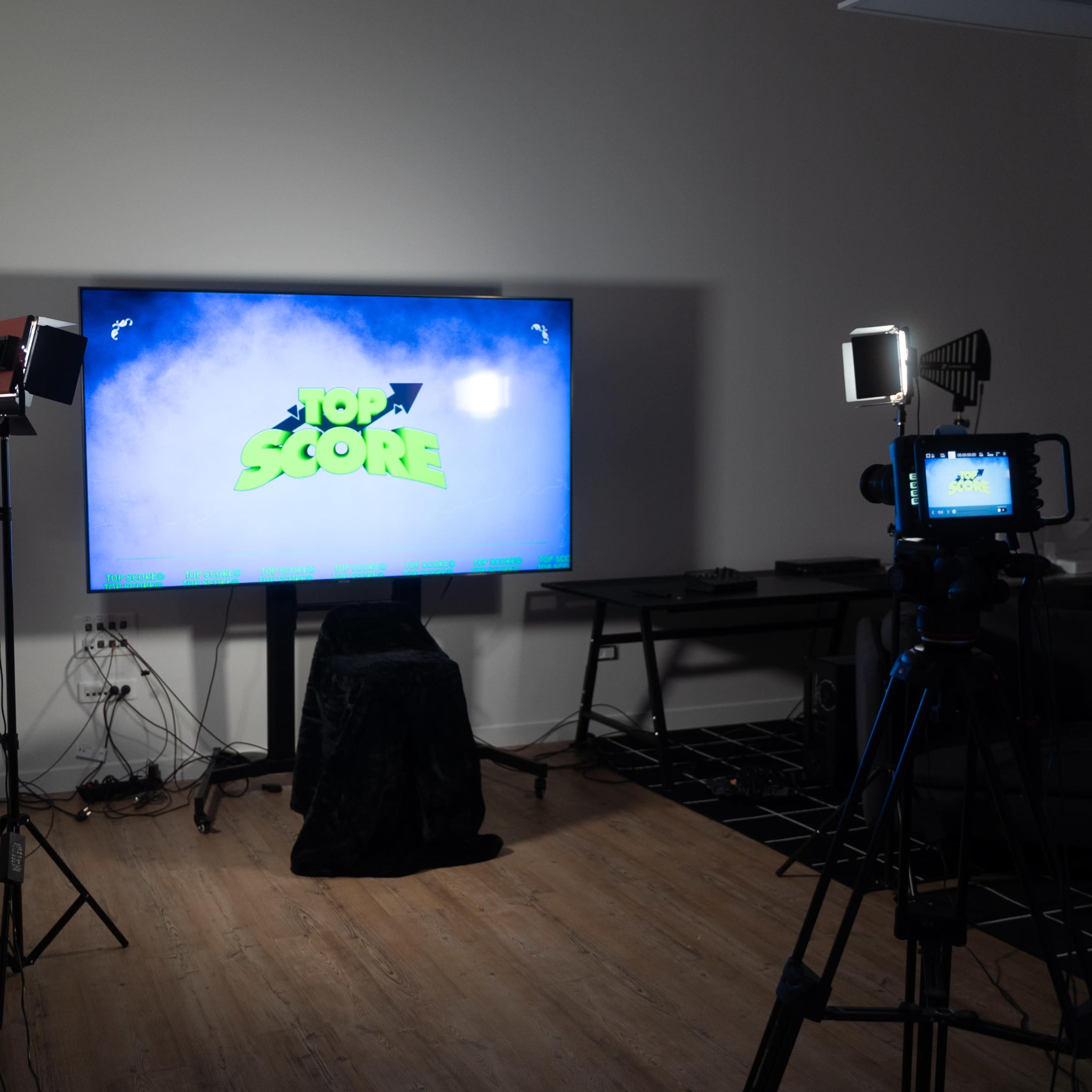 A large television displaying "TOP SCORE" is set up in a dimly lit room, accompanied by camera equipment and lighting stands.