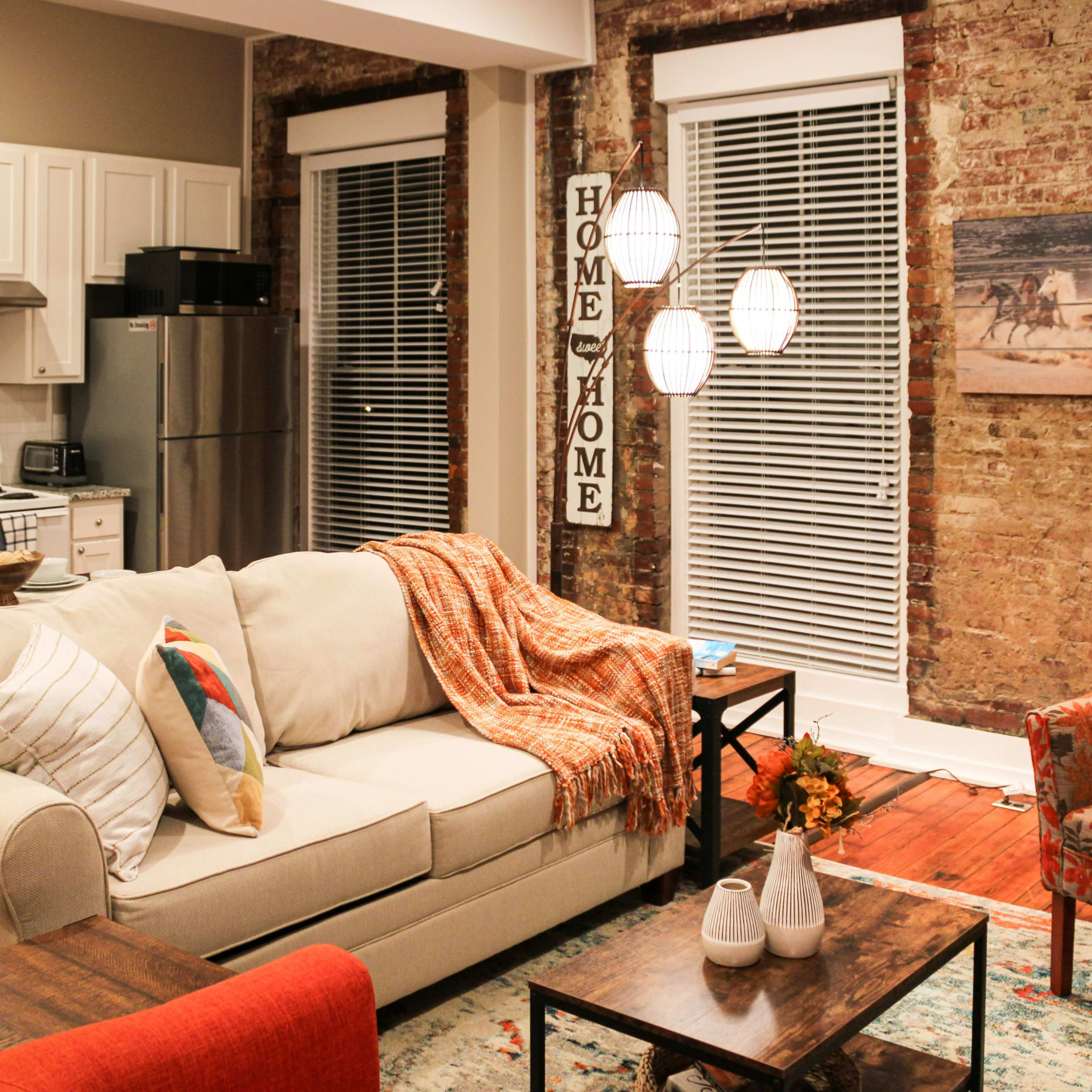 A modern living room with a light-colored sofa, a wooden coffee table, and a kitchen area in the background, featuring exposed brick walls and large windows with blinds.