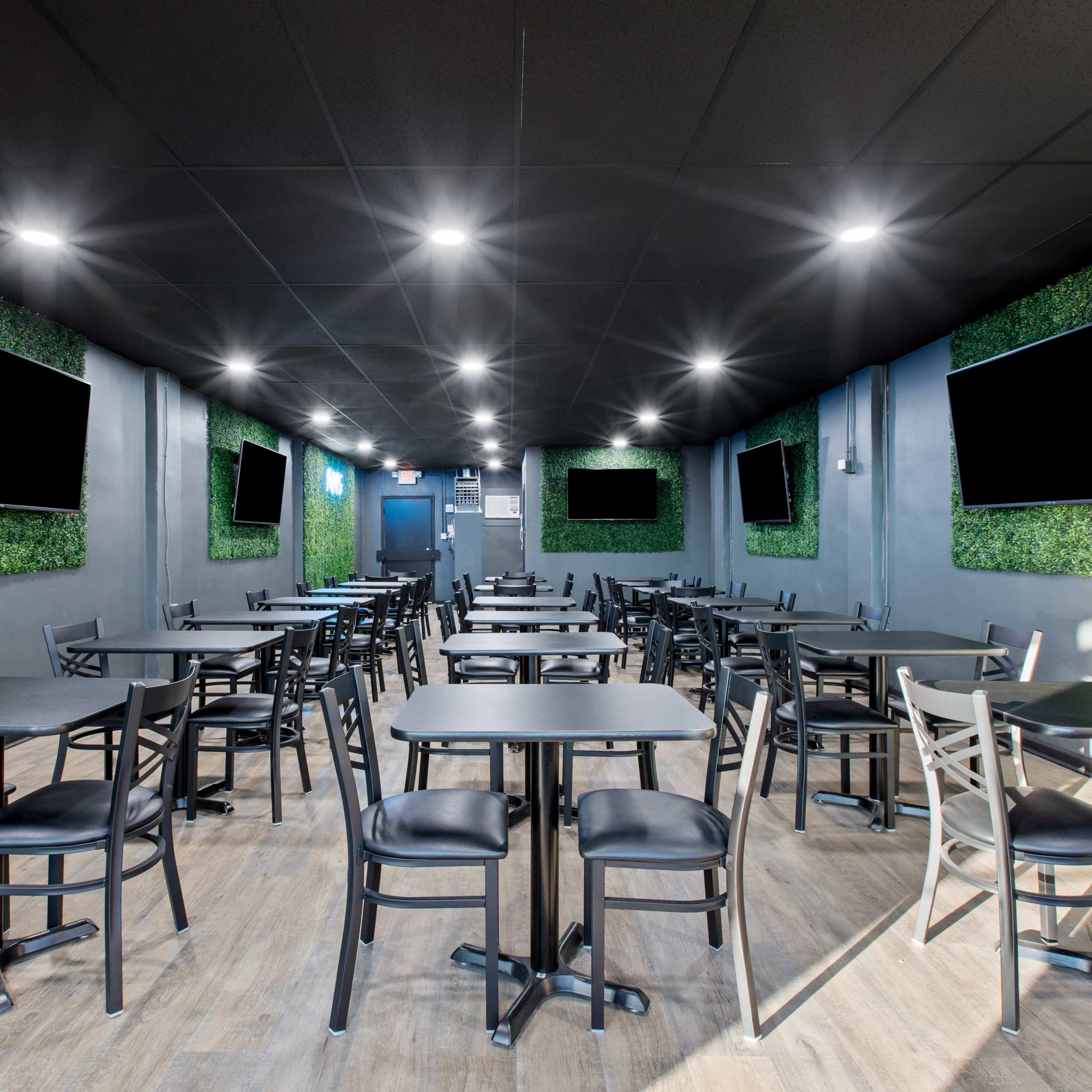 A modern restaurant interior with black tables and chairs, potted greenery on the walls, and several wall-mounted televisions.