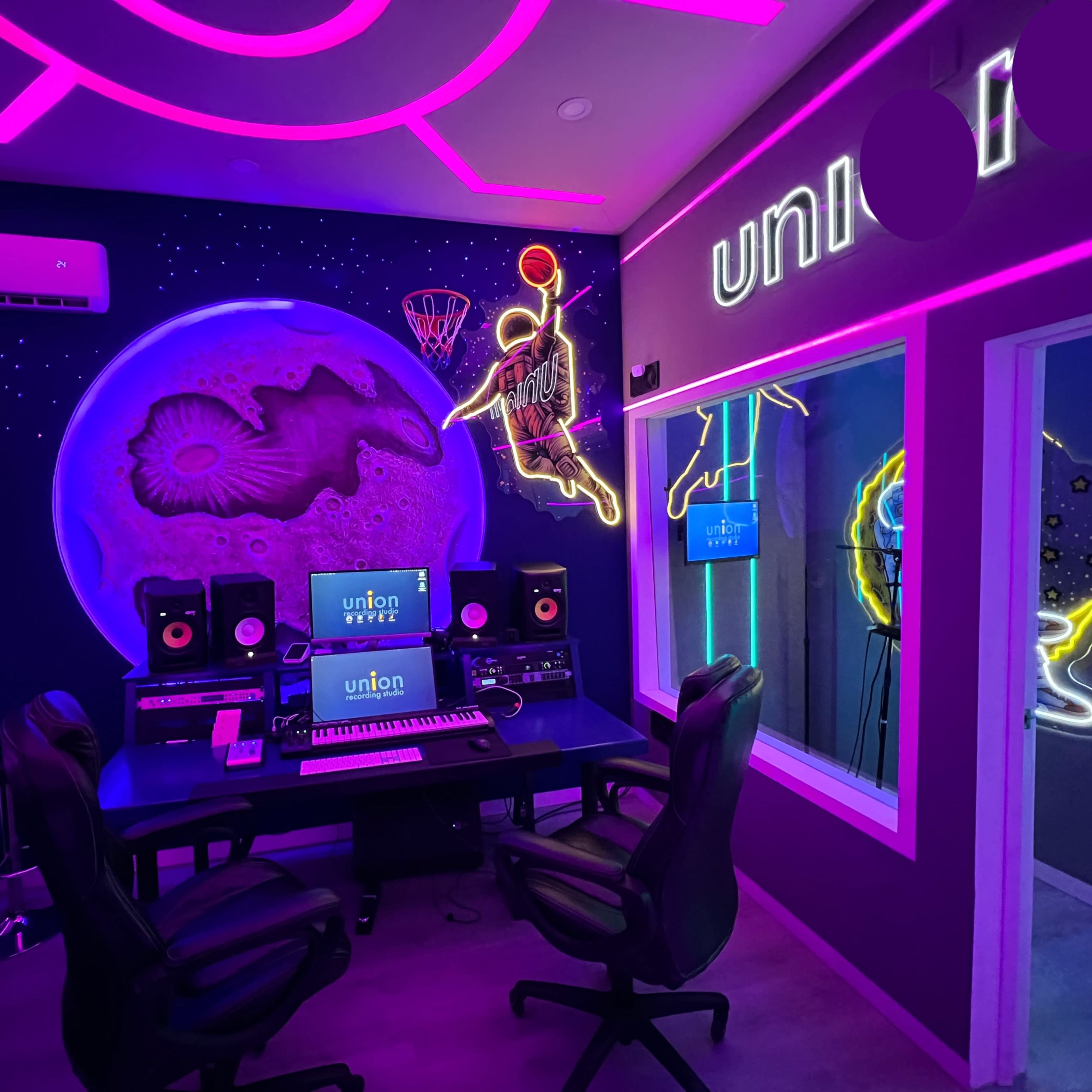 A modern music studio featuring a neon-lit interior with a large mural of a basketball player and a moon backdrop, accompanied by a central mixing desk and sound monitors.
