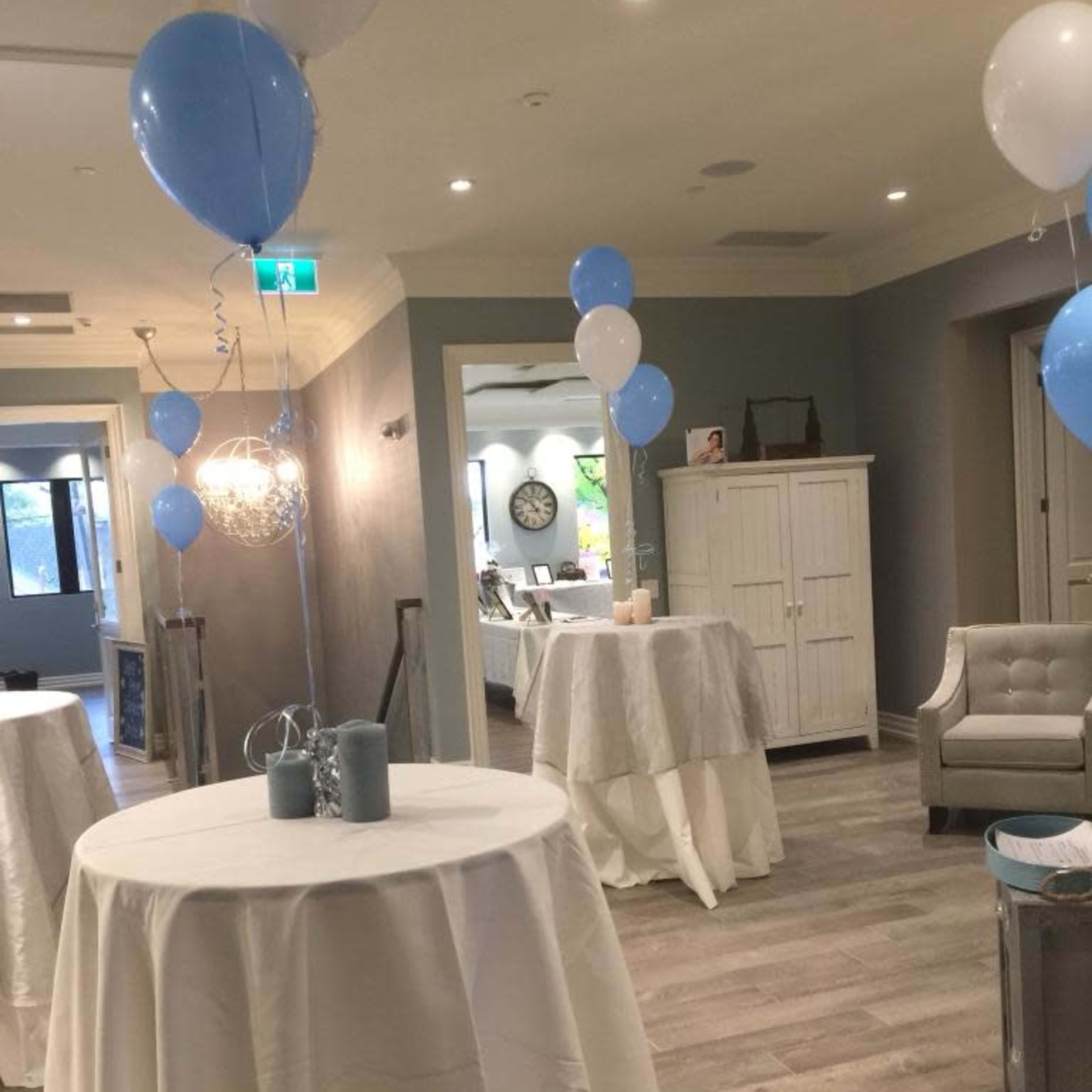 A decorated indoor space with tables covered in white tablecloths, blue and white balloons, and soft lighting.