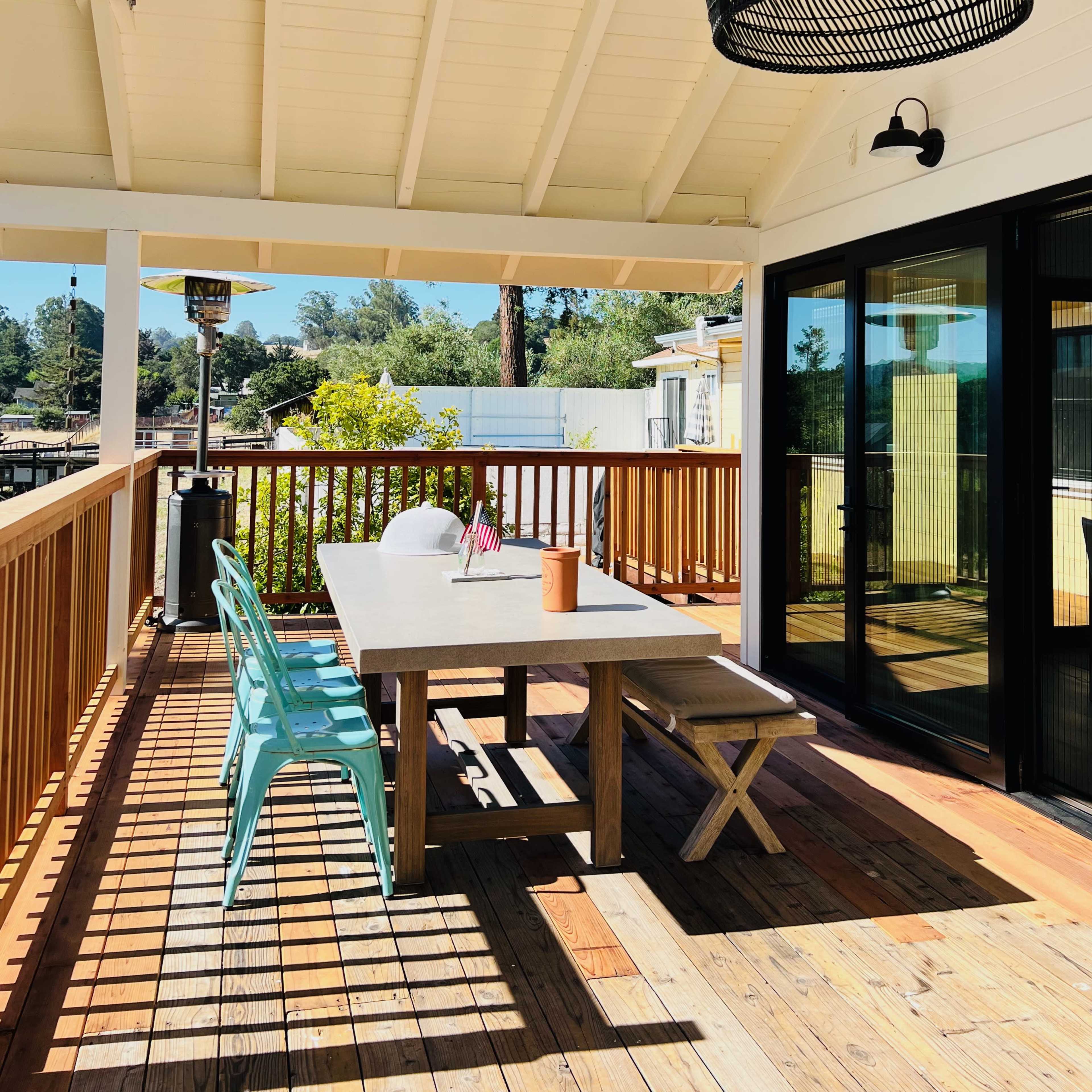 A spacious outdoor deck features a wooden table with seating, a patterned light fixture, and a view of greenery.