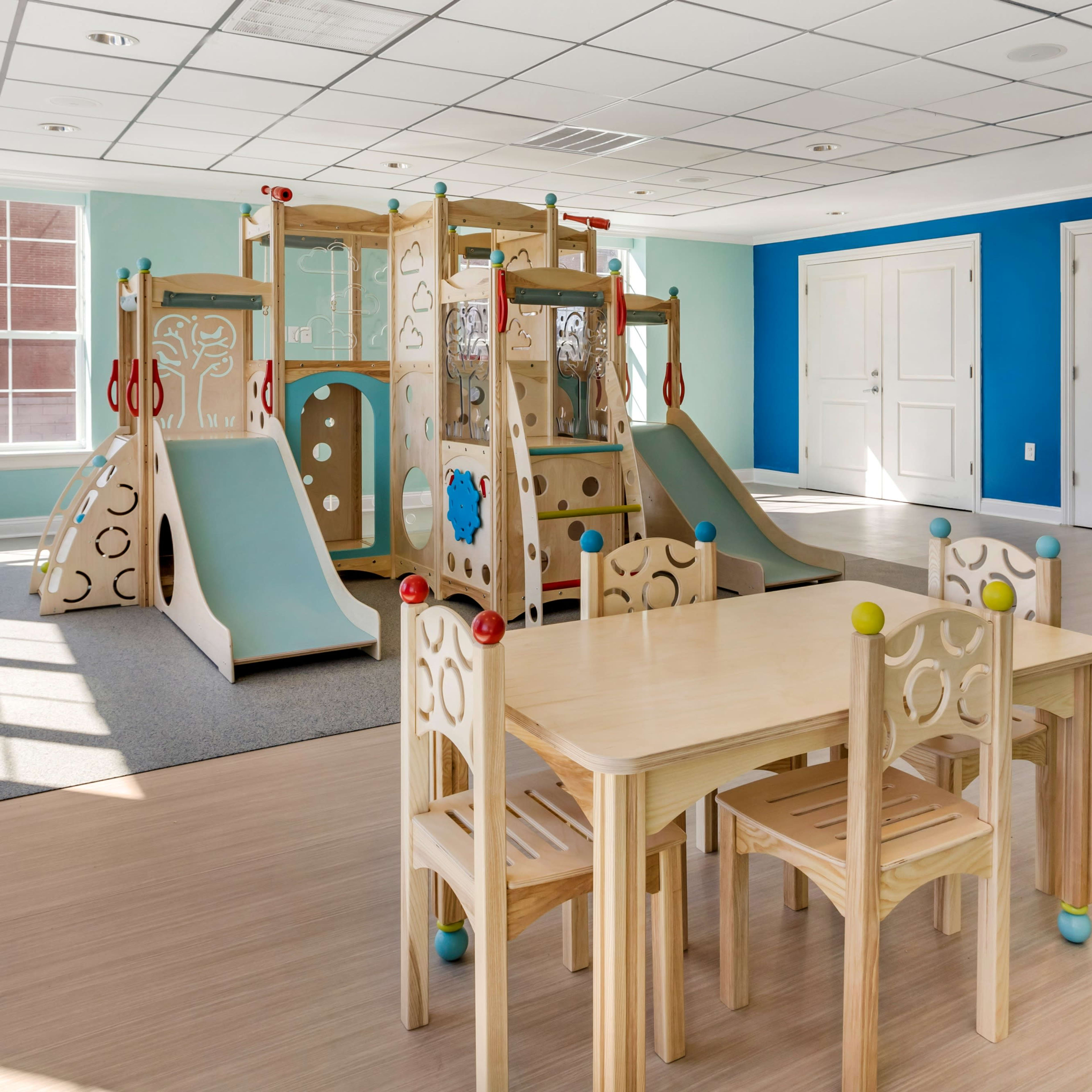 A brightly colored playroom featuring a wooden play structure and a set of three small tables with chairs.