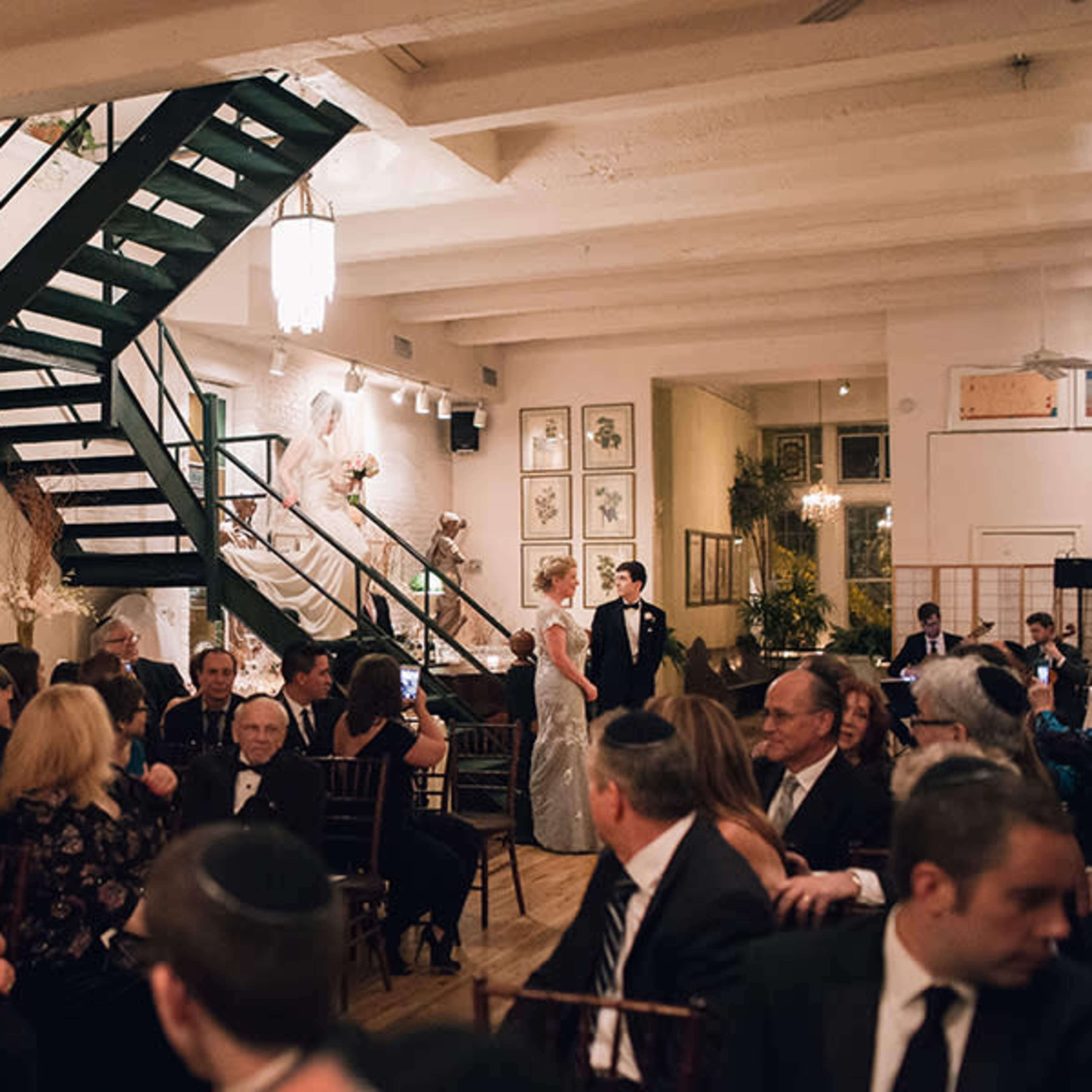 A crowded banquet hall with guests seated at tables, an elegant staircase in the background, and a couple standing near the center of the room.