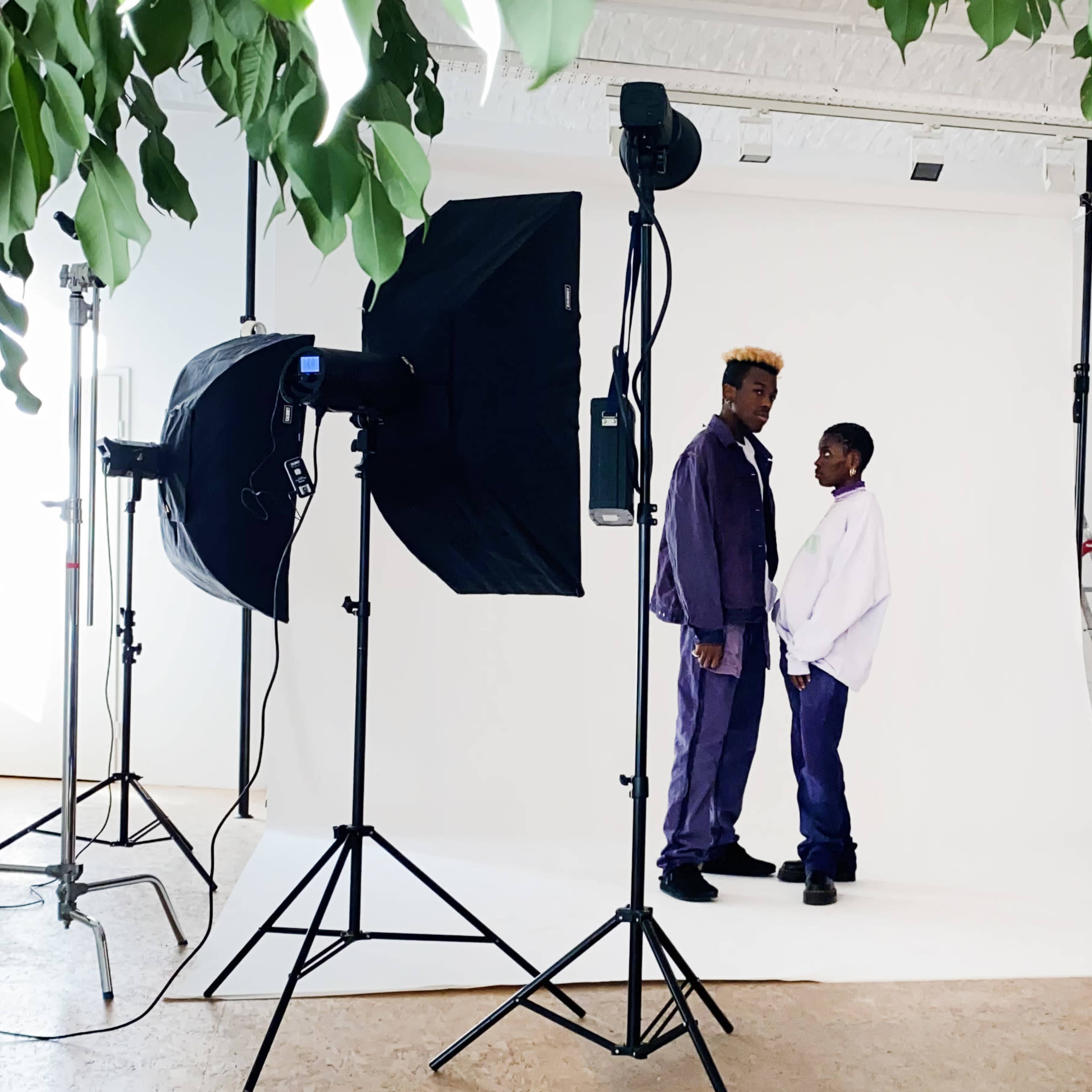 Two individuals in matching purple outfits stand in front of a white backdrop surrounded by professional photography equipment and lighting.
